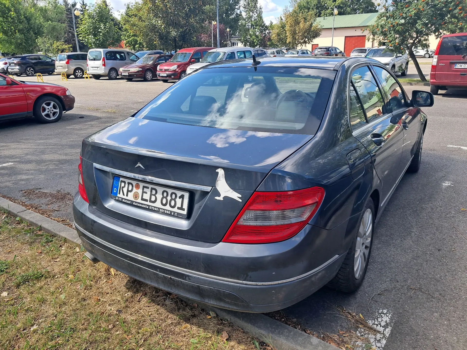 Tylny widok ciemnoniebieskiego samochodu osobowego zaparkowanego na parkingu. Na klapie bagażnika znajduje się niemiecka tablica rejestracyjna o numerze RP EB 881 oraz biała naklejka przedstawiająca sylwetkę ptaka. W tle widoczne są inne zaparkowane samochody oraz drzewa i budynek.