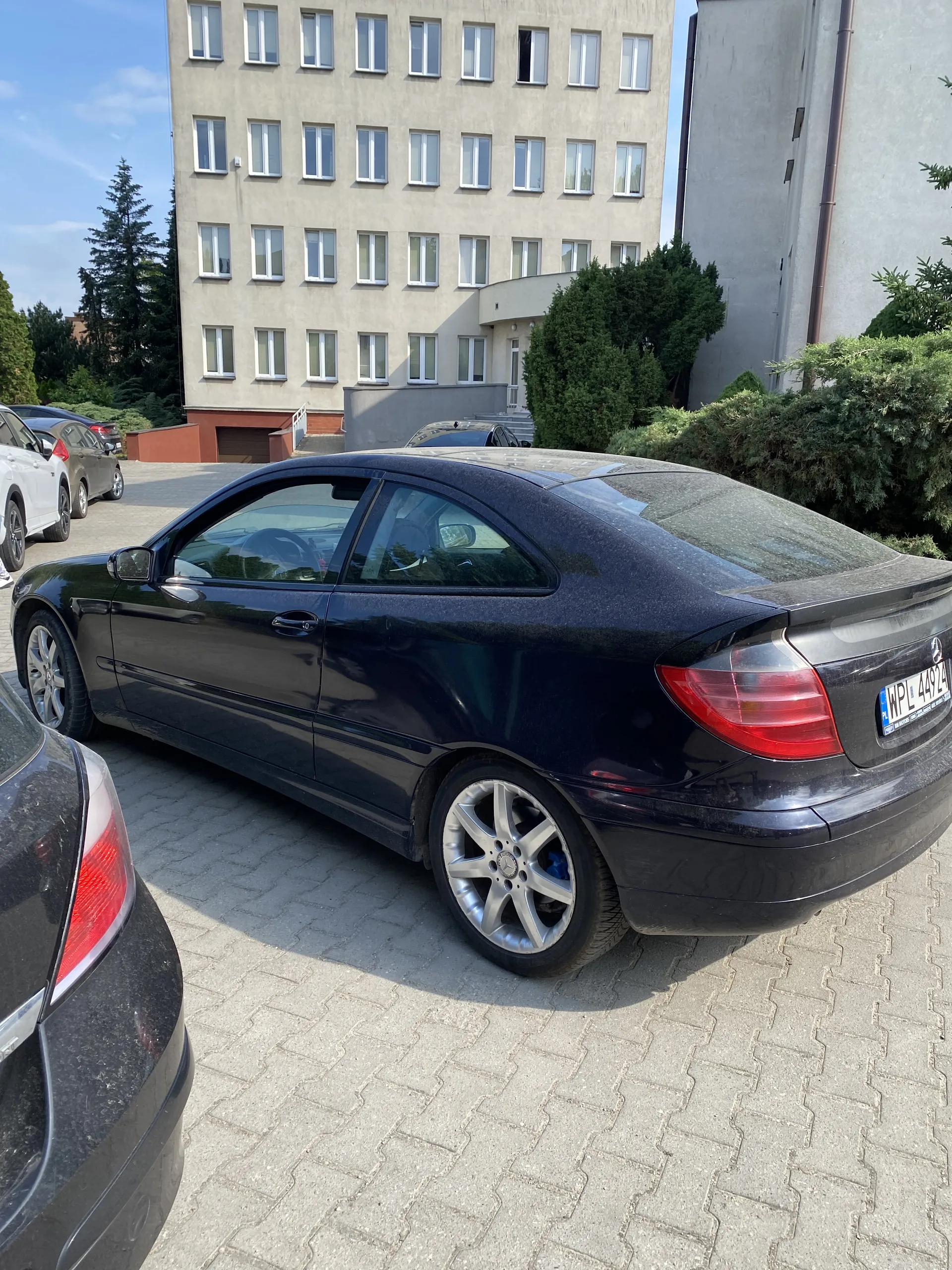 Czarny samochód osobowy zaparkowany na boku na powierzchni z kostki brukowej. Widoczna tablica rejestracyjna z napisem WPL 494L. W tle budynek wielopiętrowy z oknami ułożonymi poziomo, drzewa oraz inne zaparkowane pojazdy. Niebo jest jasne.