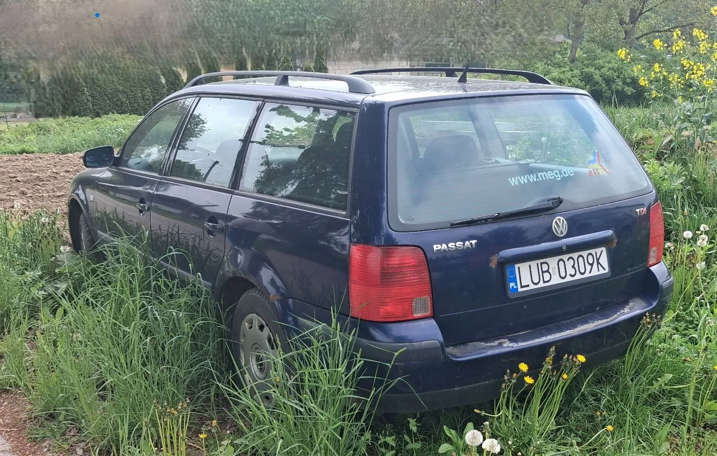 Ciemnoniebieski samochód Volkswagen Passat w nadwoziu kombi, zaparkowany w wysokiej trawie i chwastach. Na tylnej szybie znajduje się naklejka z adresem strony internetowej www.meg.de. Samochód posiada polską tablicę rejestracyjną o numerze LUB 0309K.