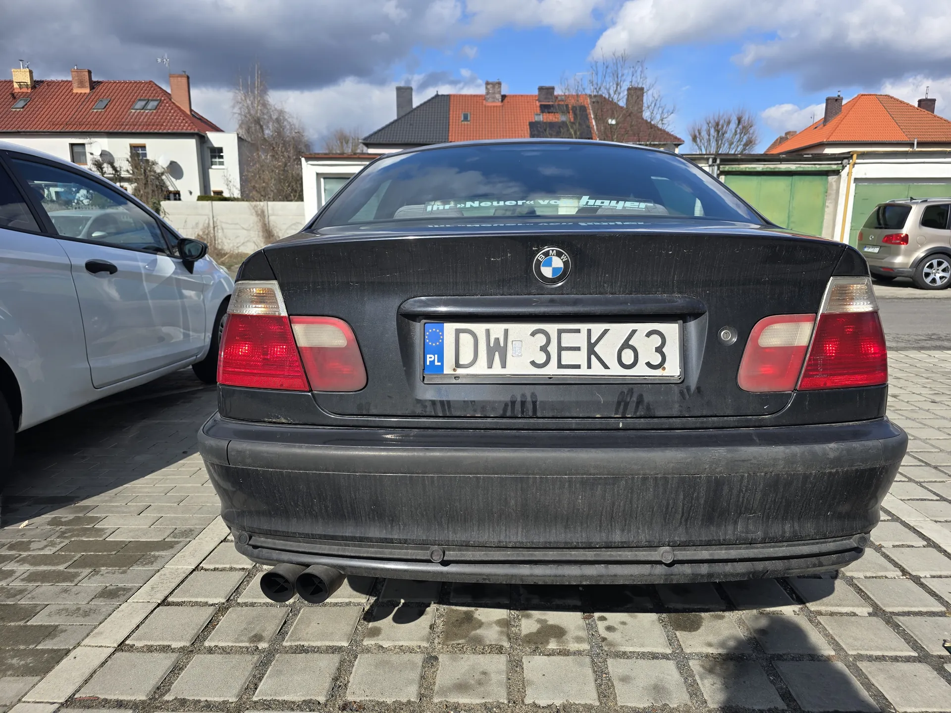 Tył czarnego samochodu marki BMW z tablicą rejestracyjną PL DW 3EK63. Po lewej stronie widoczny srebrny samochód, po prawej beżowy. Tło stanowią domy z czerwonymi dachami, drzewa i niebo z chmurami. Samochód stoi na brukowanej powierzchni. Na szybie tylnej widoczny napis: Ihr »Neuer« von hoyer.