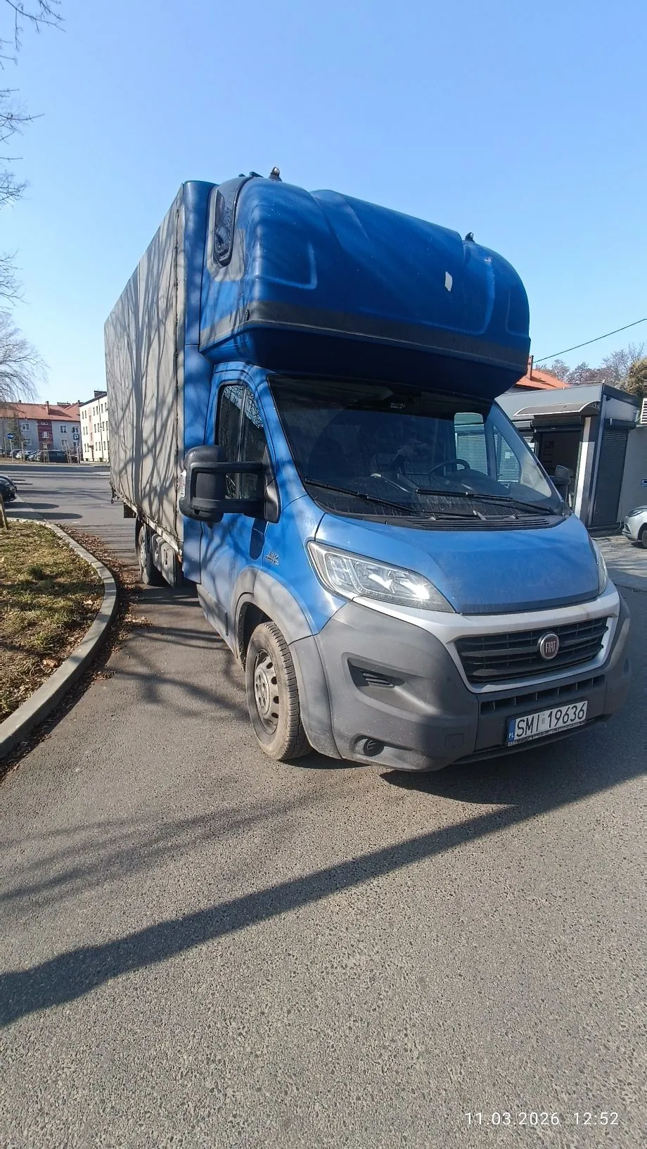 Niebieski samochód dostawczy marki Fiat z dużą nadbudową transportową, zaparkowany na poboczu asfaltowej drogi. Pojazd posiada tablicę rejestracyjną o numerze SMI 19636. W tle widoczne są niskie budynki mieszkalne pod bezchmurnym, niebieskim niebem. W prawym dolnym rogu zdjęcia znajduje się data i godzina: 11.03.2026 12:52.