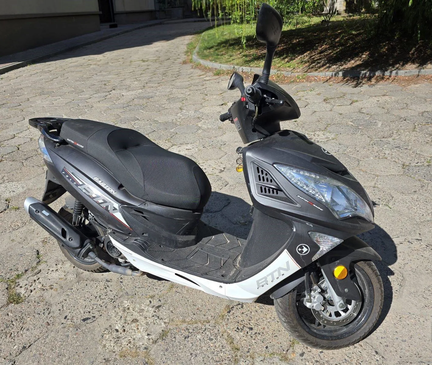 Czarny skuter marki Barton, model Falcon, zaparkowany na brukowanej drodze. Pojazd ma czarną karoserię z białymi elementami na dolnej części ramy. Na boku widnieje napis 'BARTON' oraz 'FALCON'. Skuter jest wyposażony w przedni reflektor, lusterka boczne i czarny wydech.