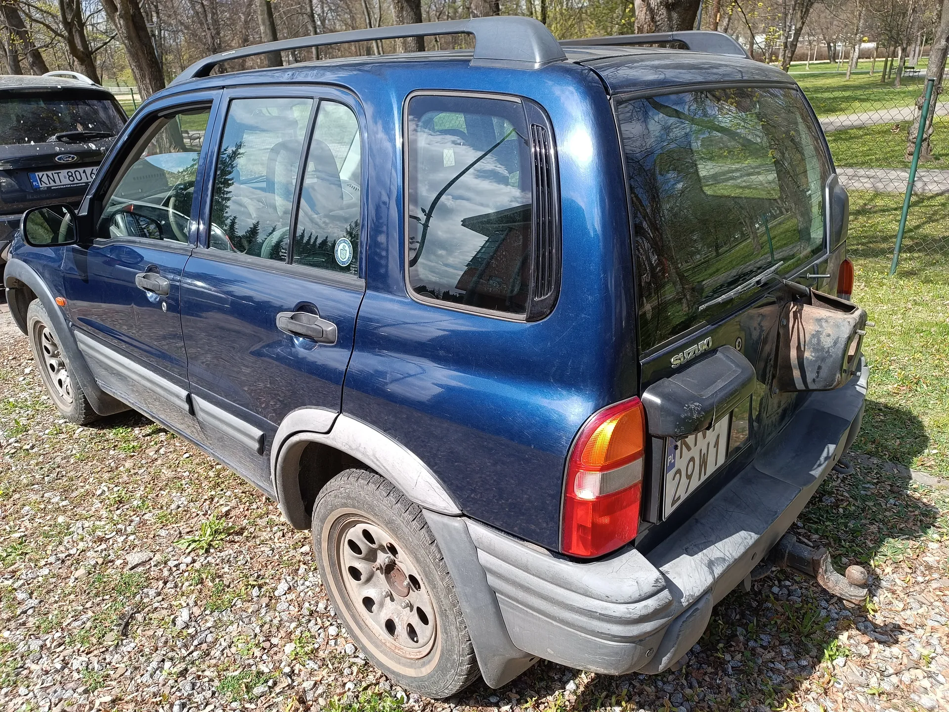 Niebieski samochód typu SUV marki Suzuki, widok z tyłu i boku. Widoczna tablica rejestracyjna PL 29W1, dach z przenośnymi barami, tylne światła oraz oznaczenie marki na bagażniku. Samochód stoi na podłożu z żwiru i trawy, w tle drzewa, ogrodzenie i inny pojazd z tablicą KNT 8016. W oknach odbijają się chmury i budynki.