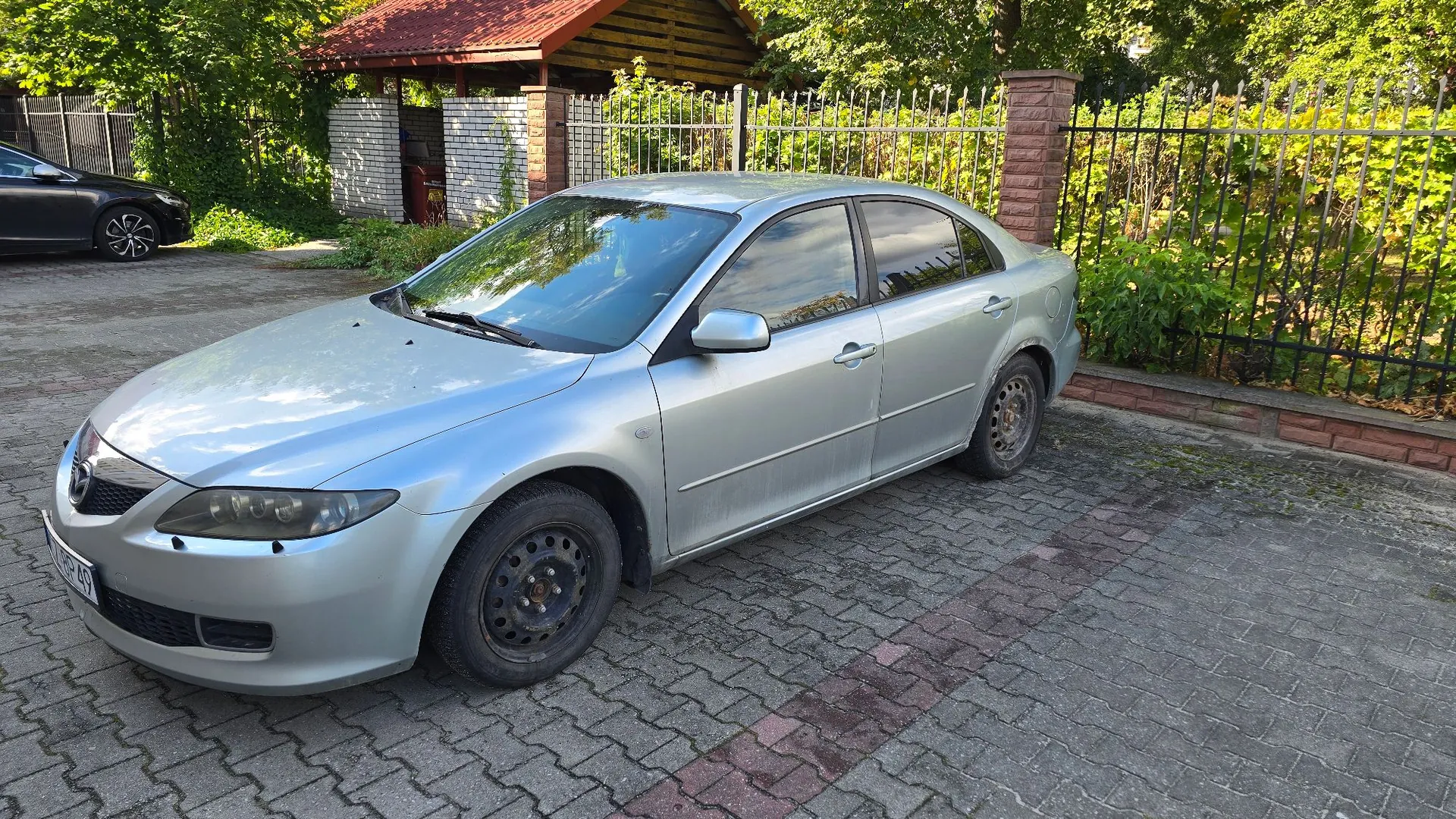 Srebrny samochód osobowy typu sedan zaparkowany na podjeździe z kostki brukowej. Auto jest ustawione pod kątem, widoczny jest przód oraz prawy bok pojazdu. W tle znajduje się metalowy płot, zielone krzewy, drzewa oraz fragment drewnianego budynku z czerwonym dachem.