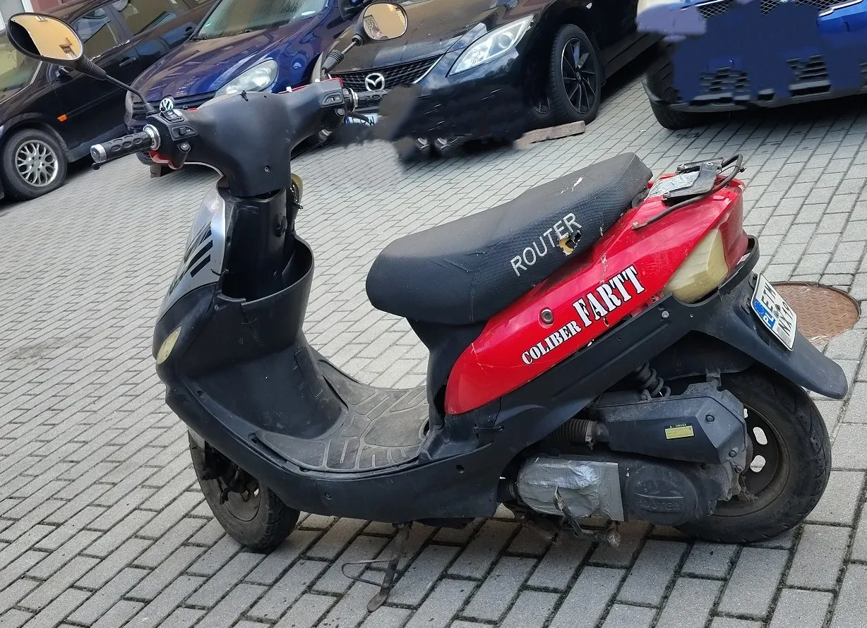 Czarny skuter z czerwonym tylnym panelem, zaparkowany na kostce brukowej. Na czerwonej części widnieje biały napis „ROUTER” oraz „COLIBER FARTTT”. W tle widoczne są zaparkowane samochody w kolorach czarnym i niebieskim.