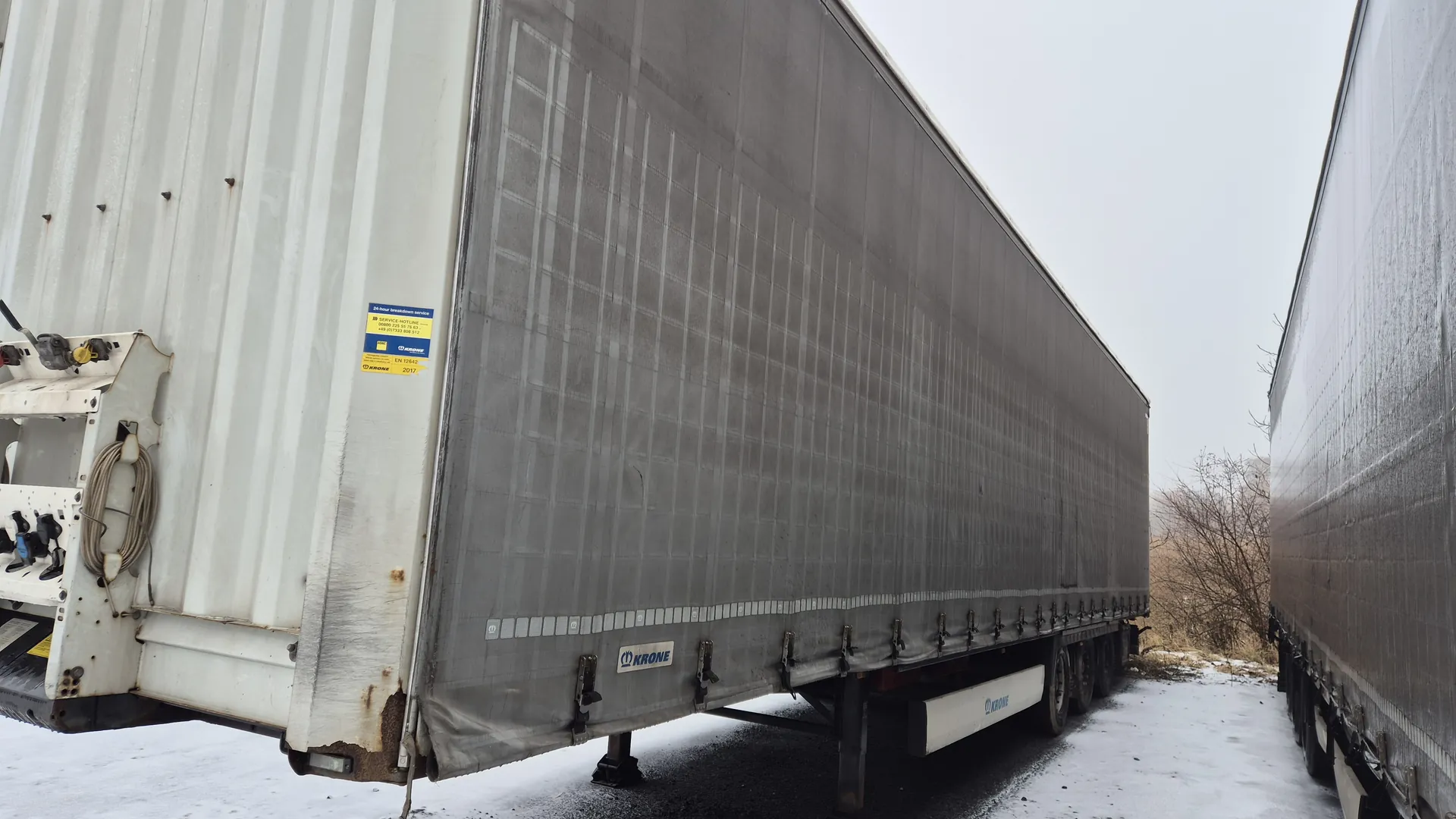 Duża przyczepa ciężarowa z szarą tkaniną pokrywającą ładunek, biały kontener po lewej stronie z niebiesko-żółtymi naklejkami. Widoczny napis 'KRONE' na tkaninie i białym elemencie przyczepy. Na tle druga przyczepa, śnieg na ziemi, suche krzewy i pochmurne niebo.