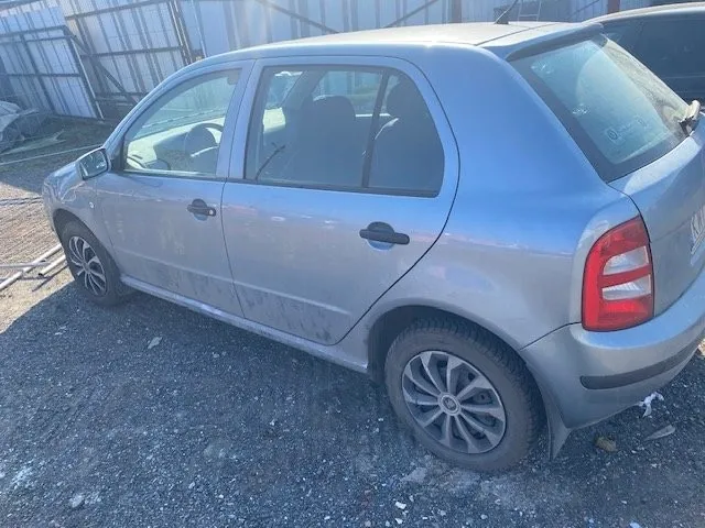 Srebrny samochód osobowy typu hatchback z czterema drzwiami, zaparkowany na żwirowym podłożu. Samochód ma zabrudzone boki, ciemne szyby i felgi z wieloramiennym wzorem. W tle widoczna metalowa konstrukcja budynku oraz fragmenty innych pojazdów. Podłoże pokryte jest drobnymi kamieniami i śmieciem.