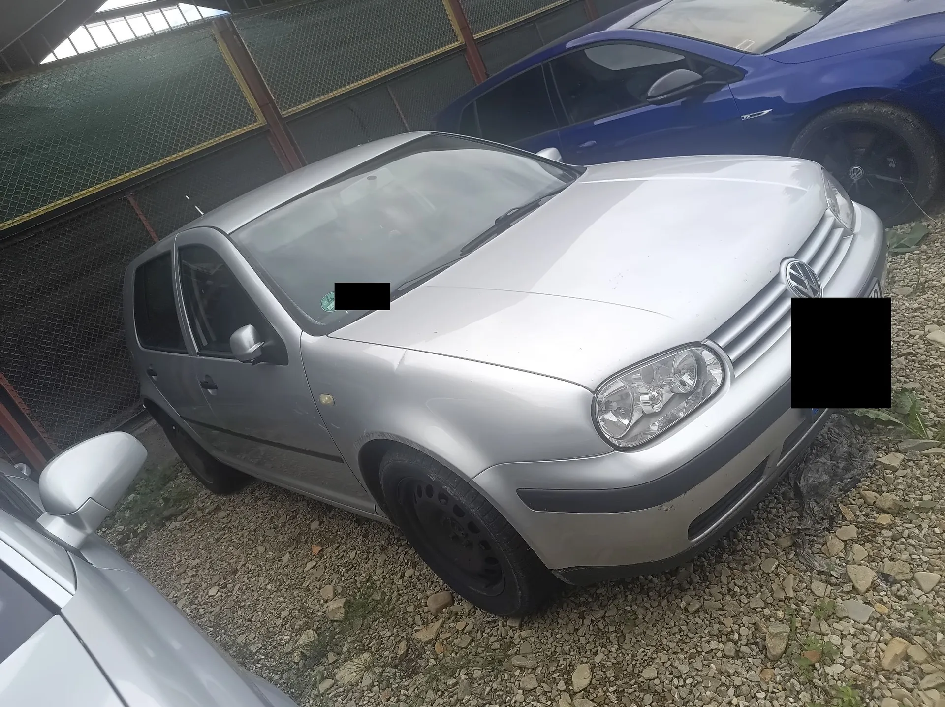 Srebrny samochód osobowy marki Volkswagen Golf zaparkowany na żwirowym podłożu. Obok niego widoczny jest fragment niebieskiego samochodu oraz fragment innego srebrnego pojazdu w lewym dolnym rogu. W tle znajduje się ogrodzenie z siatki.