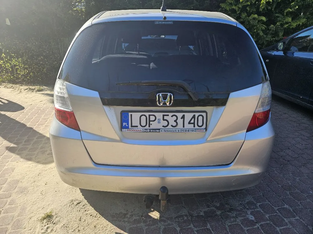 Zdjęcie przedstawia tył srebrnego samochodu marki Honda z tablicą rejestracyjną PL LOP 53140. Na tablicy widoczny jest napis "HONDA" oraz dodatkowy tekst pod tablicą. Samochód stoi na brukowanej powierzchni, w tle zielone krzewy i fragment czarnego pojazdu po prawej stronie. Słoneczne oświetlenie rzuca cienie na ziemię.