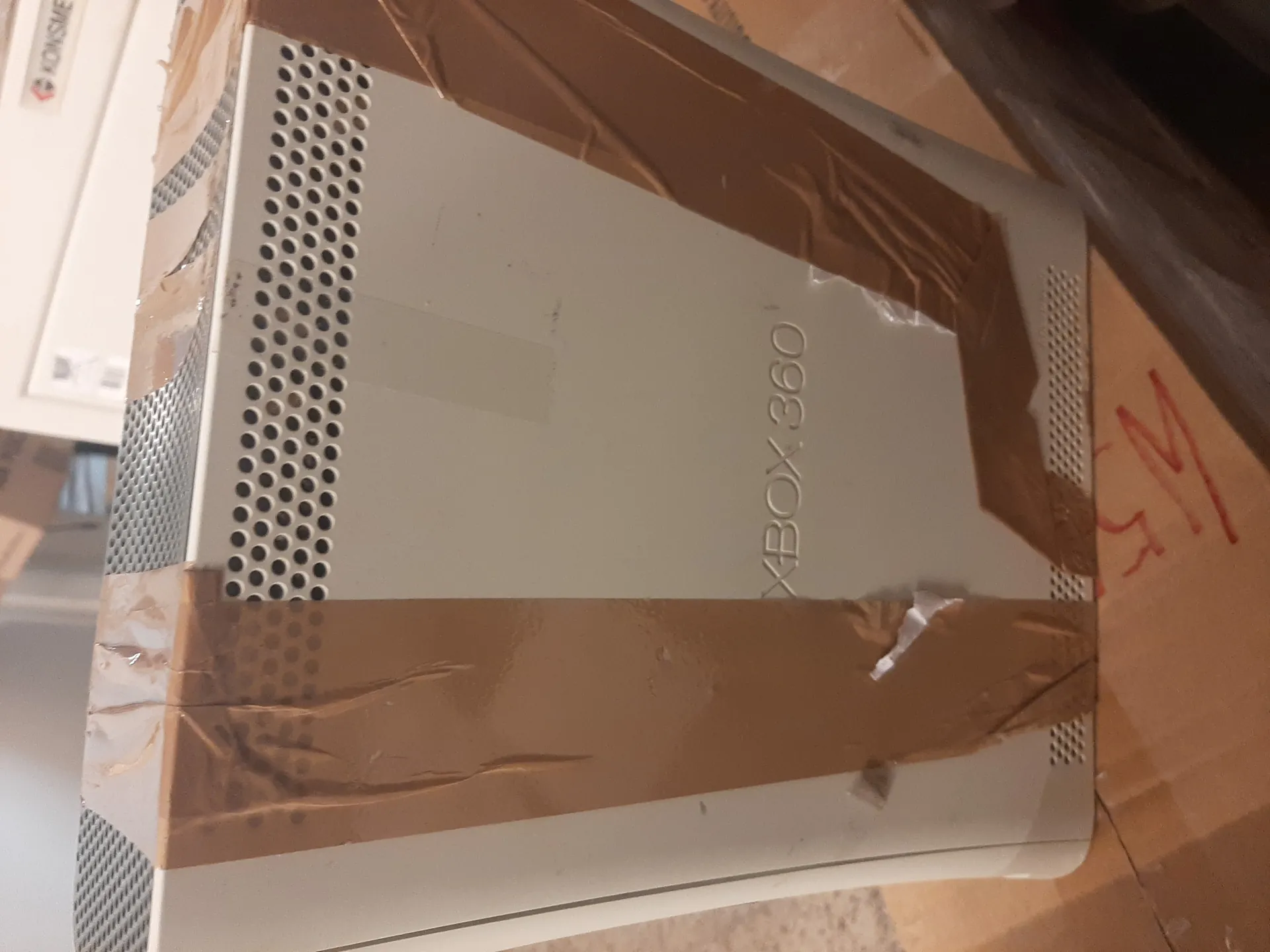 Biały konsolowy aparat Xbox 360 z widocznym napisem "XBOX 360" na przedniej części. Urządzenie jest owinięte brązową taśmą klejącą, która częściowo uszkodzona. Na dole obrazu widoczny jest fragment kartonu z czerwonymi oznaczeniami. W tle znajduje się biały obiekt z częściowo widocznym napisem "KONISME".