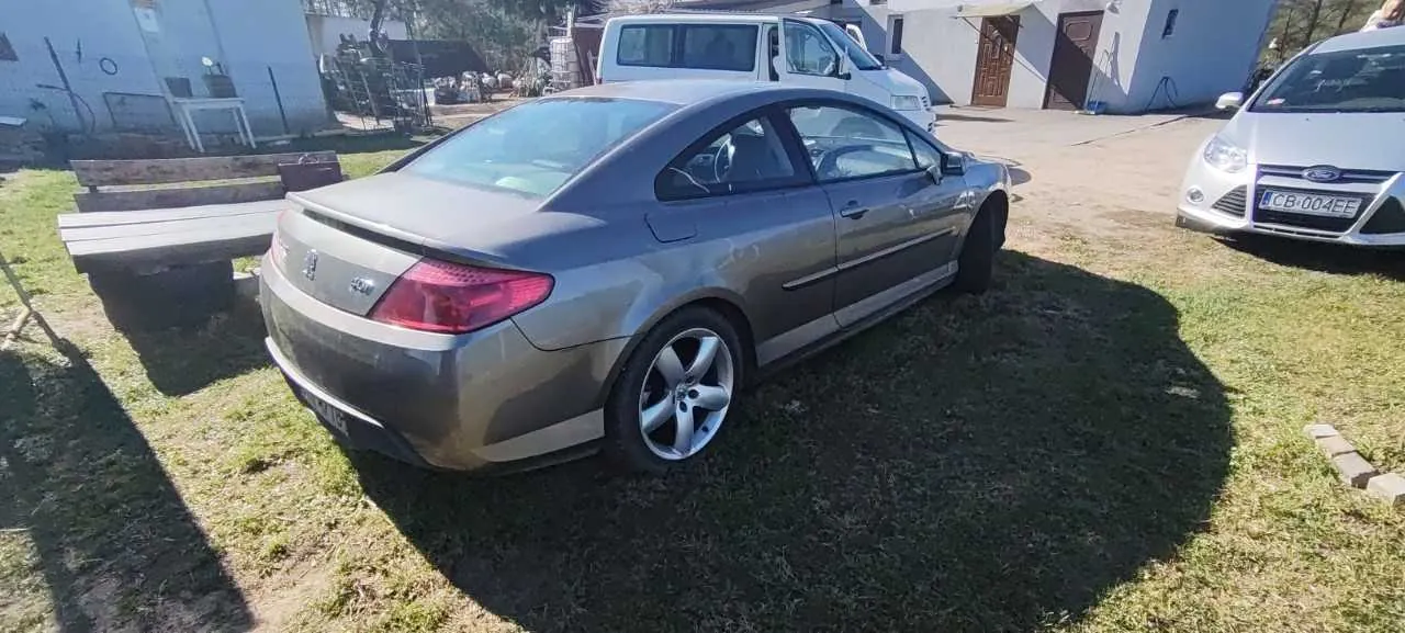 Srebrny samochód osobowy typu sedan zaparkowany na trawie, widok z tyłu i boku. W tle biały samochód z czytelną tablicą rejestracyjną CB-004EE oraz biały van. Widoczne są budynki, ławka i trawa. Po prawej stronie częściowo widoczny srebrny samochód marki Ford.