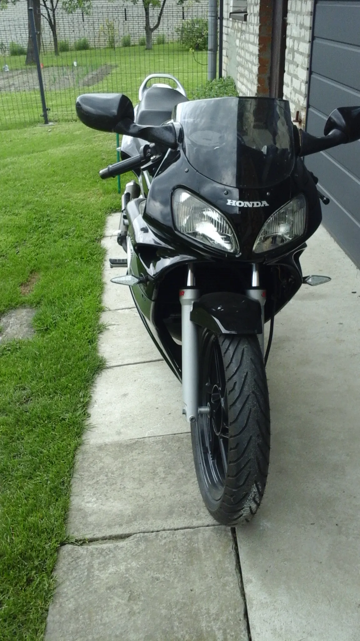 Czarny motocykl marki Honda z dwoma reflektorami, widok z przodu. Na masce widoczny pionowy napis HONDA. Motocykl umieszczony poziomo na betonowej nawierzchni, w tle trawa, ogrodzenie i budynek.
