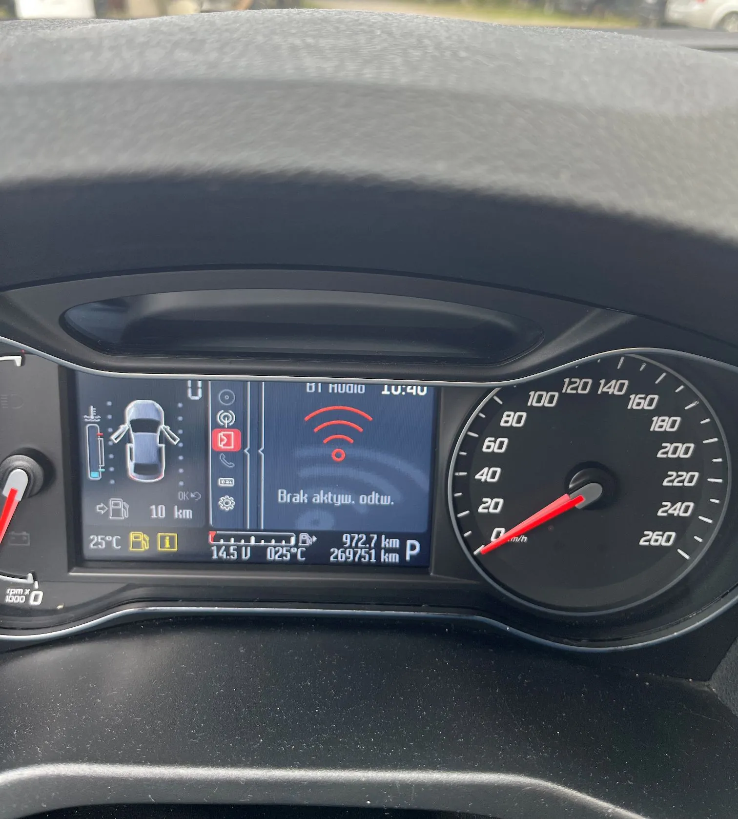 Zdjęcie deski rozdzielczej samochodu. Po prawej stronie znajduje się analogowy prędkościomierz z czerwoną wskazówką wskazującą 0 km/h. W centrum znajduje się cyfrowy wyświetlacz z informacjami: godzina 10:40, napis 'BT AUDIO', ikona fal radiowych oraz komunikat 'Brak aktyw. odtw.'. Wyświetlacz pokazuje również schemat samochodu z otwartymi drzwiami, temperaturę zewnętrzną 25°C, napięcie akumulatora 14.5 V, temperaturę oleju 025°C, zasięg 972.7 km oraz całkowity przebieg 269751 km. Wybrany jest bieg P.
