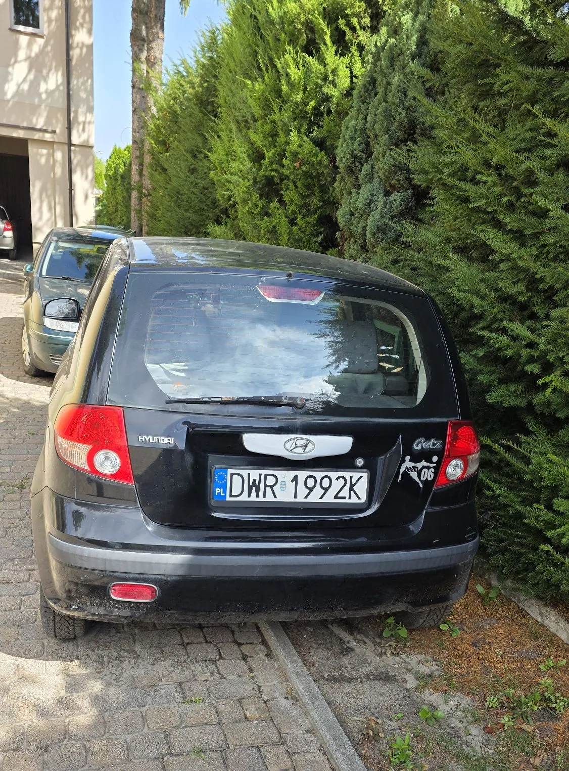 Tylny widok czarnego samochodu marki Hyundai Getz zaparkowanego na kostce brukowej obok zielonego żywopłotu. Samochód ma polską tablicę rejestracyjną o numerze DWR 1992K. W tle widoczny jest fragment jasnego budynku oraz inny, zielony samochód.