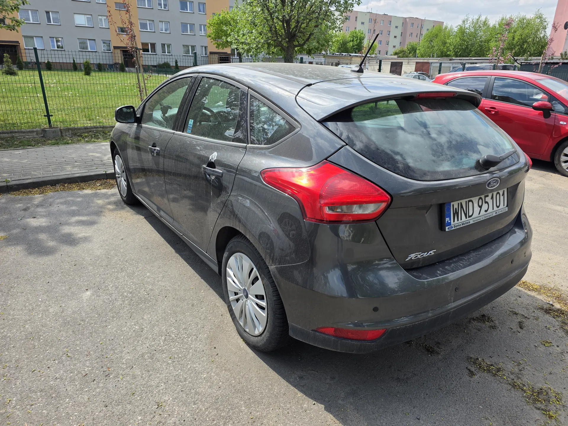 Szary samochód Ford Focus z tablicą rejestracyjną WND 95101 zaparkowany na asfaltowym placu. W tle czerwony samochód, trawa za ogrodzeniem, bloki mieszkalne, drzewa i częściowo zachmurzone niebo. Na tylnym zderzaku widoczny napis Focus oraz logo Forda.