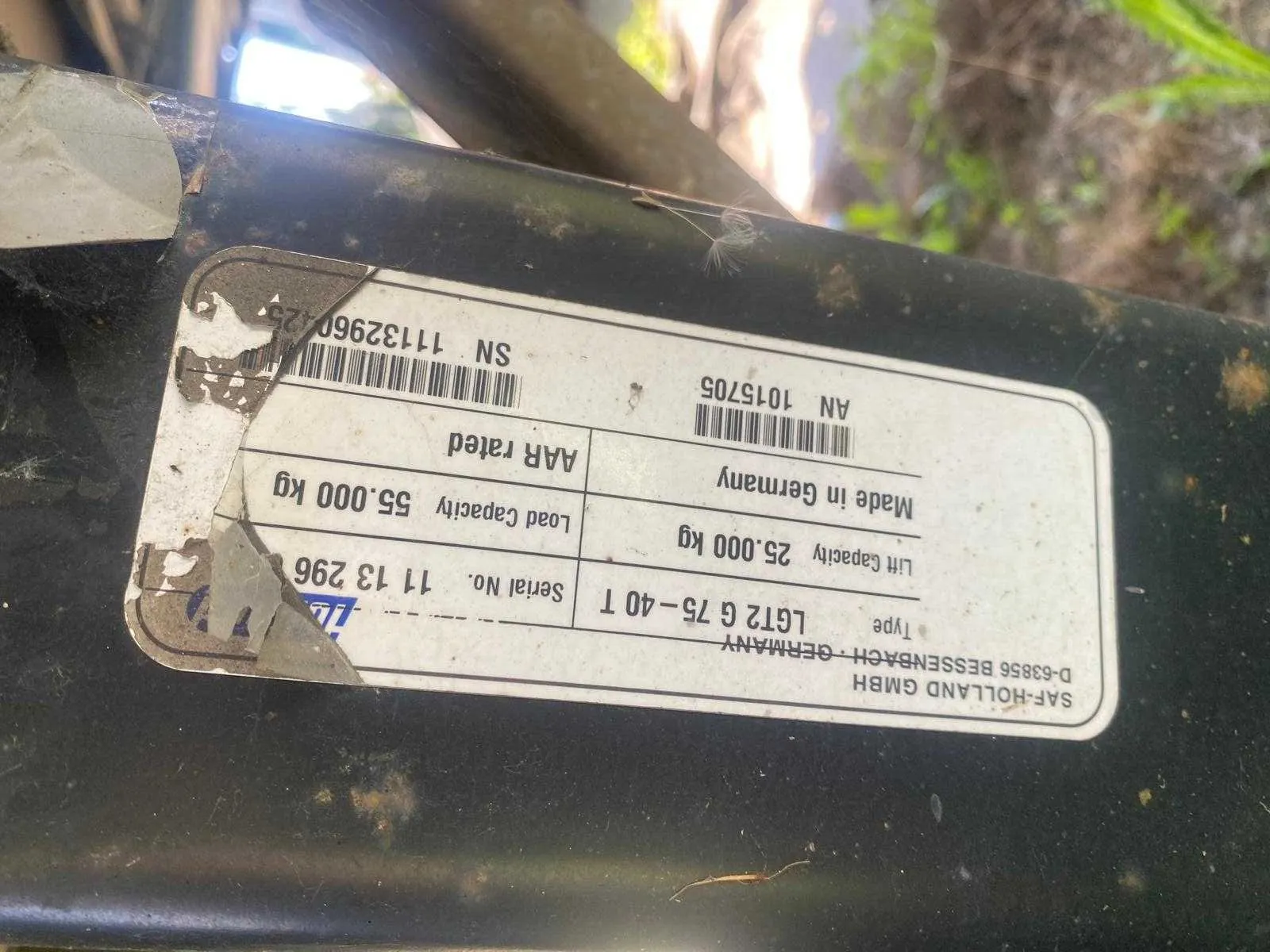 Zdjęcie tabliczki znamionowej przymocowanej do metalowej ramy pojazdu. Tabliczka zawiera następujące informacje: producent SAF-HOLLAND GMBH, adres D-63856 BESSENBACH - GERMANY, typ LGT2 G 75-40 T, Lift Capacity 25.000 kg, Made in Germany, Serial No. 11 13 296, Load Capacity 55.000 kg, AAR rated oraz numer seryjny SN 11132960 z kodem kreskowym i numerem AN 1015705 z kodem kreskowym.