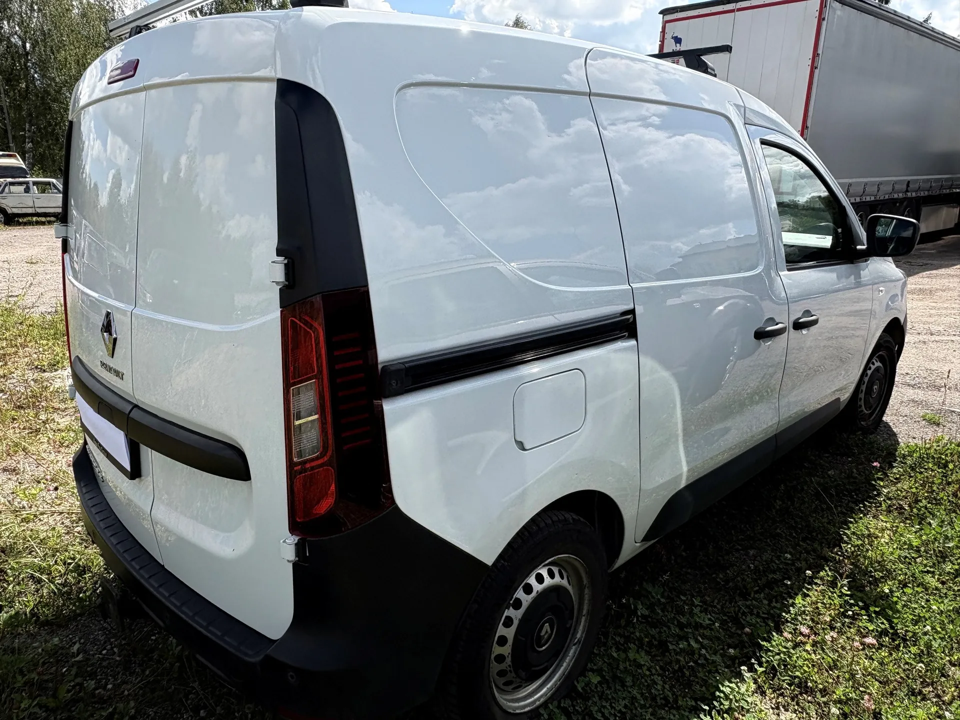 Biały samochód dostawczy marki Renault, widok z tyłu i z boku. Pojazd posiada czarne plastikowe elementy wykończenia z tyłu i z boku, a także czarne felgi stalowe. W tle widoczny jest fragment białej naczepy ciężarówki oraz zielona trawa.
