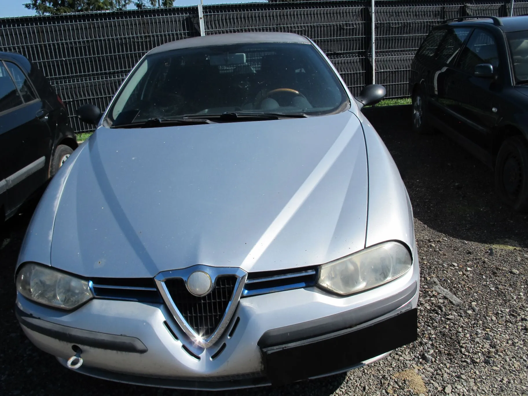 Srebrny samochód marki Alfa Romeo zaparkowany przodem do obiektywu na żwirowym podłożu. Po lewej i prawej stronie widoczne są fragmenty innych, czarnych pojazdów. W tle znajduje się wysoki, ciemny płot z metalowych prętów.