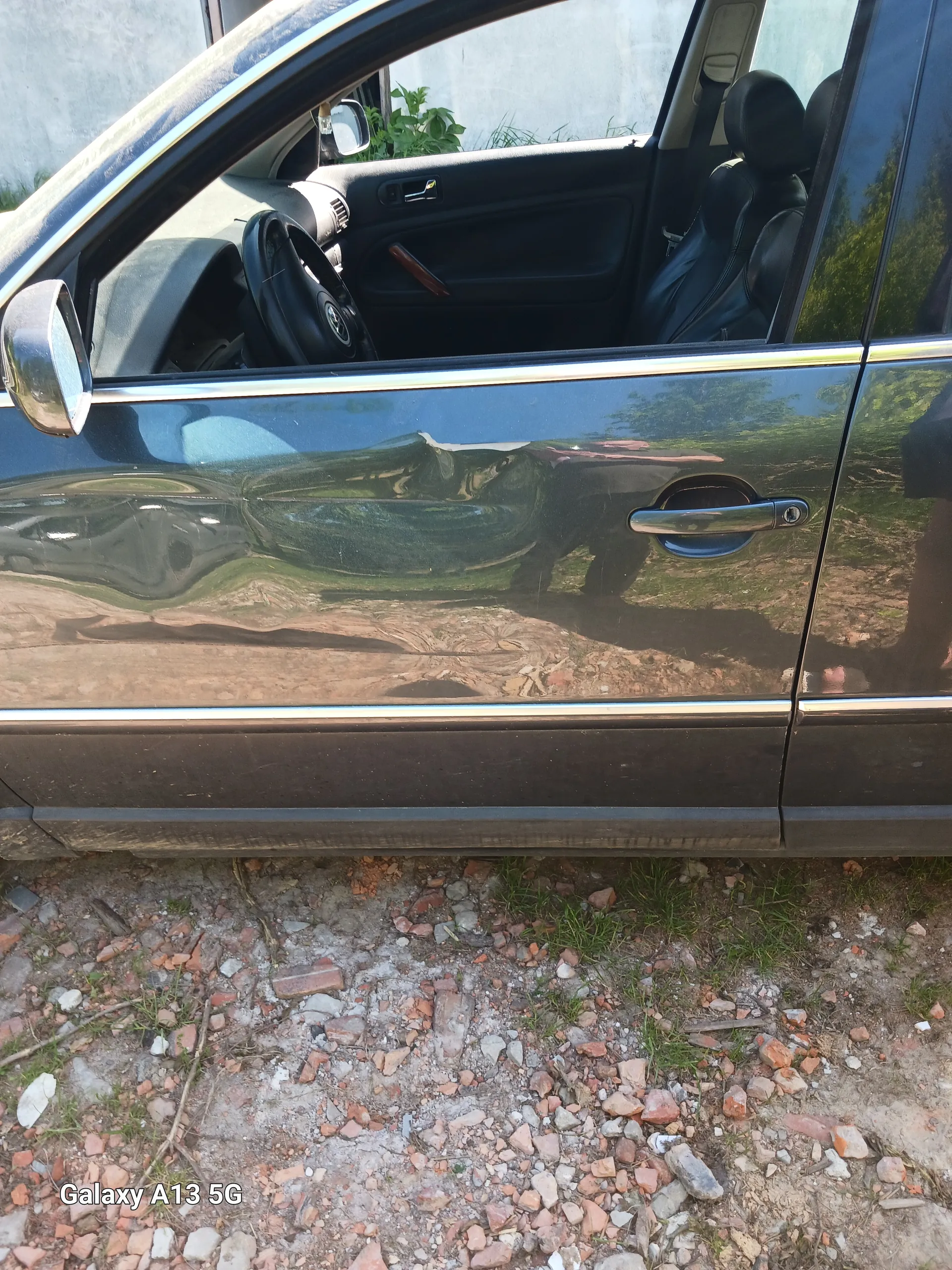 Czarny samochód z otwartą przednią lewą drzwiami. Widoczne wnętrze z kierownicą, czarnymi fotelami i fragmentem deski rozdzielczej. Zewnętrzna część drzwi z klamką i lusterkiem bocznym. Odbicie drzew i osoby w błyszczącej powierzchni drzwi. Po prawej stronie obrazu ziemia z drobno rozsypanymi cegiełkami, kamieniami i trawą. W prawym dolnym rogu widoczny pionowy napis 'Galaxy A13 5G'.