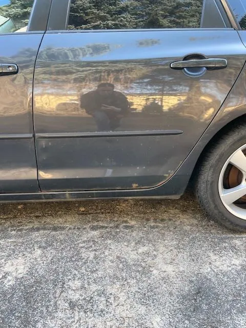 Boczny widok szarego samochodu osobowego, obejmujący drzwi z uchwytem, fragment koła tylnego oraz odbicie osoby trzymającej urządzenie w lustrzanym błyszczącym nadwoziu. W tle widoczne są drzewa i ogrodzenie. Samochód stoi na asfaltowej powierzchni z widocznymi pęknięciami i plamami.