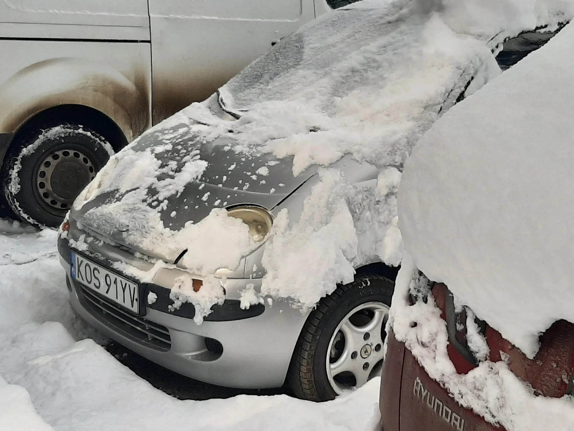 Trzy samochody pokryte grubą warstwą śniegu. W centrum srebrny samochód z widoczną tablicą rejestracyjną KOS 91YV, częściowo zakrytą śniegiem. Po lewej stronie biały furgon z brudnymi plamami na boku, po prawej czerwony samochód marki Hyundai z widocznym napisem na masce. Śnieg pokrywa całą powierzchnię pojazdów i ziemię wokół.