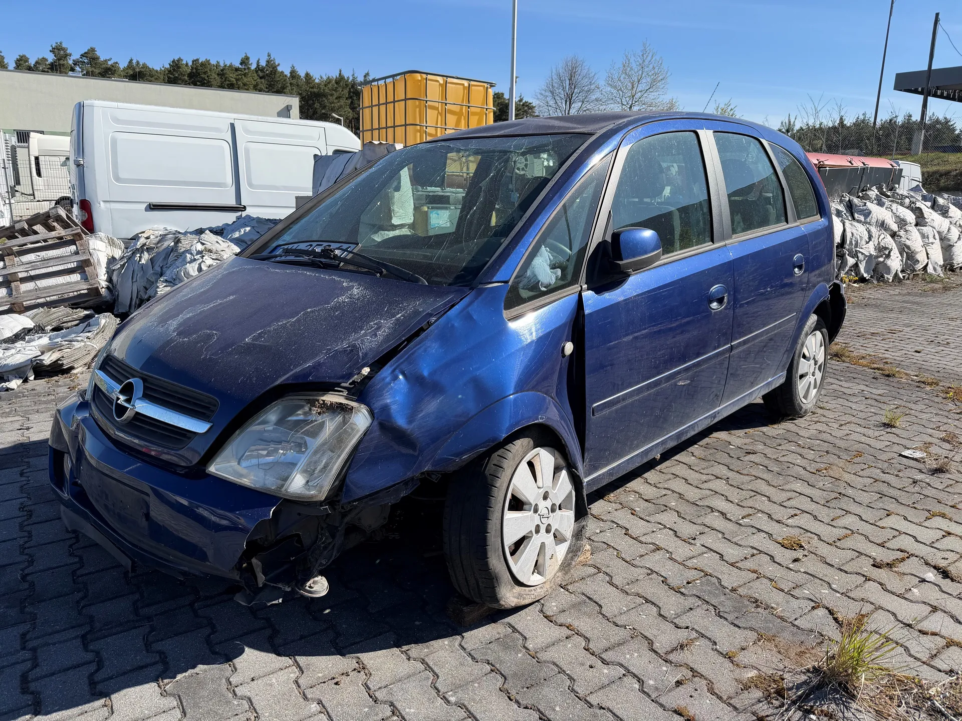 Niebieski samochód osobowy z uszkodzonym przodem, w tym zdeformowaną maską, pękniętym reflektorem i pękniętym przednim szybą. Samochód stoi na brukowanej powierzchni. W tle widoczny biały furgon, żółty zbiornik, stosy białych worków, drzewa oraz budynek. Niebo jest bezchmurne.