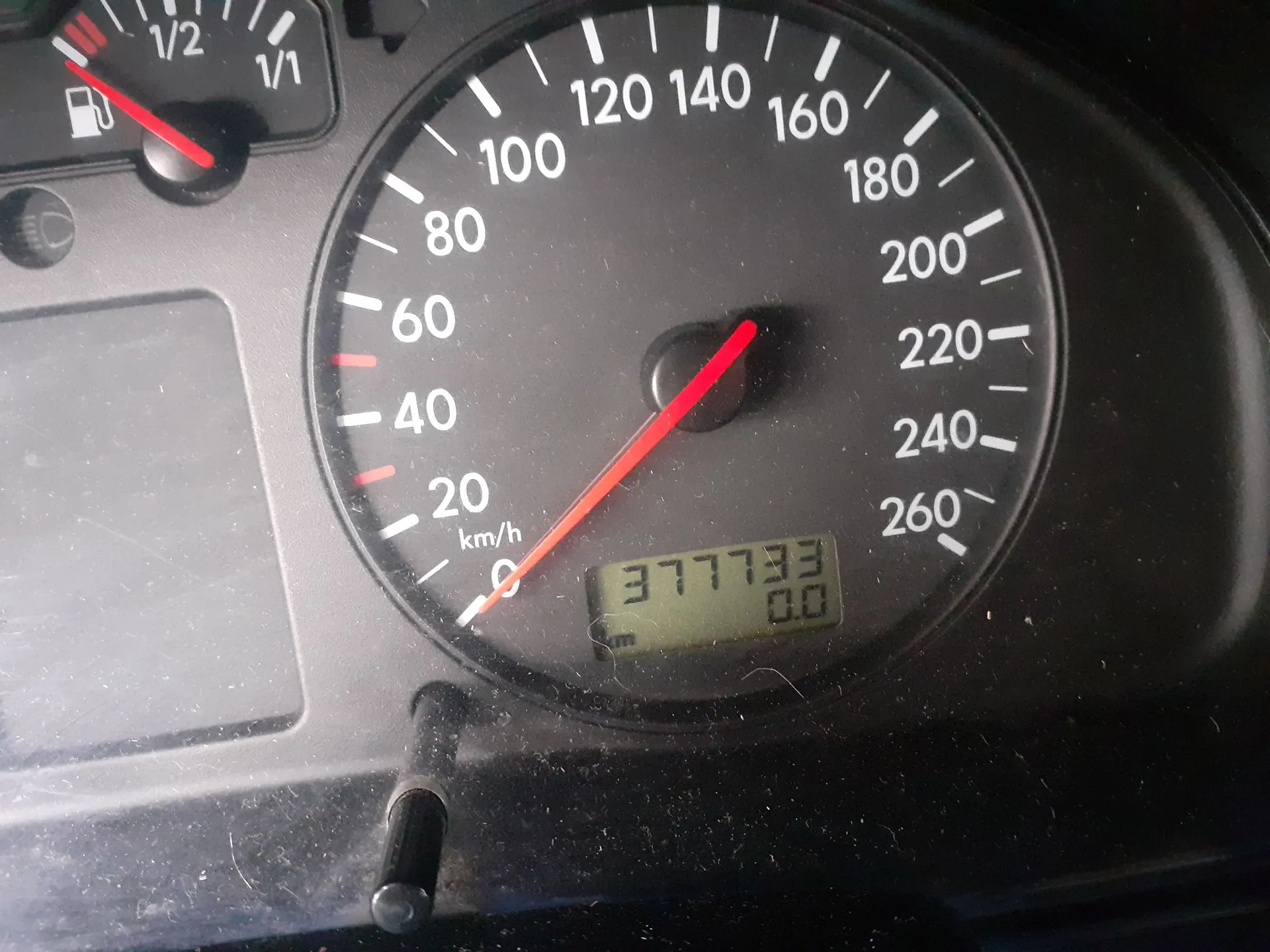 Zbliżenie na prędkościomierz w samochodzie. Wskazówka znajduje się w pozycji zero. Tarcza ma skalę od 0 do 260 km/h. Na dole znajduje się cyfrowy wyświetlacz z przebiegiem 377733 km oraz wartością 0.0. W lewym górnym rogu widoczny jest fragment wskaźnika poziomu paliwa.