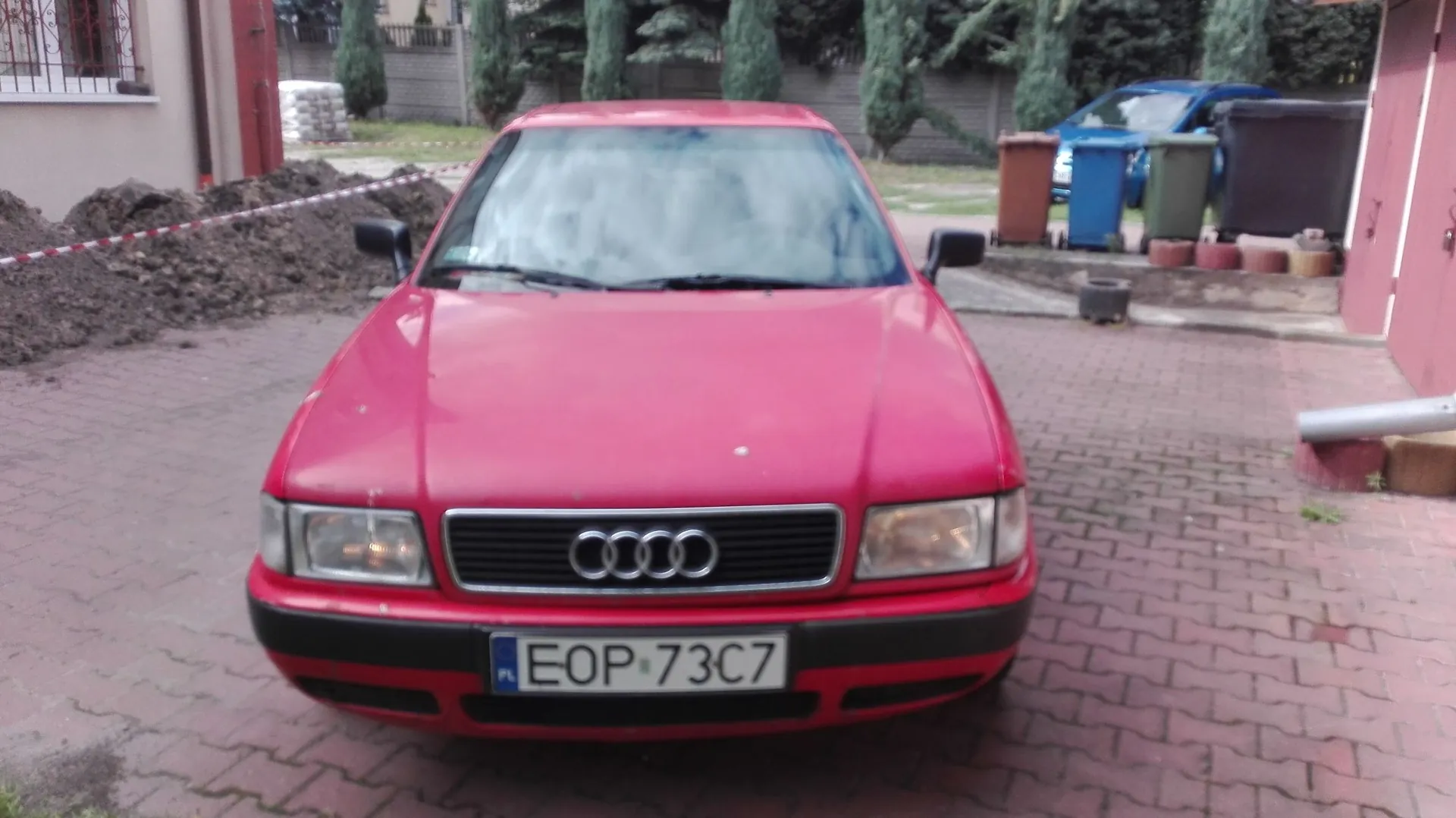 Czerwony samochód marki Audi zaparkowany przodem do obiektywu na kostce brukowej. Na przodzie auta widoczny jest grill z logo czterech pierścieni oraz tablica rejestracyjna o numerze EOP 73C7. W tle widać zielone krzewy, niebieski samochód, brązowe i niebieskie pojemniki na odpady oraz stertę ziemi odgrodzoną biało-czerwoną taśmą.