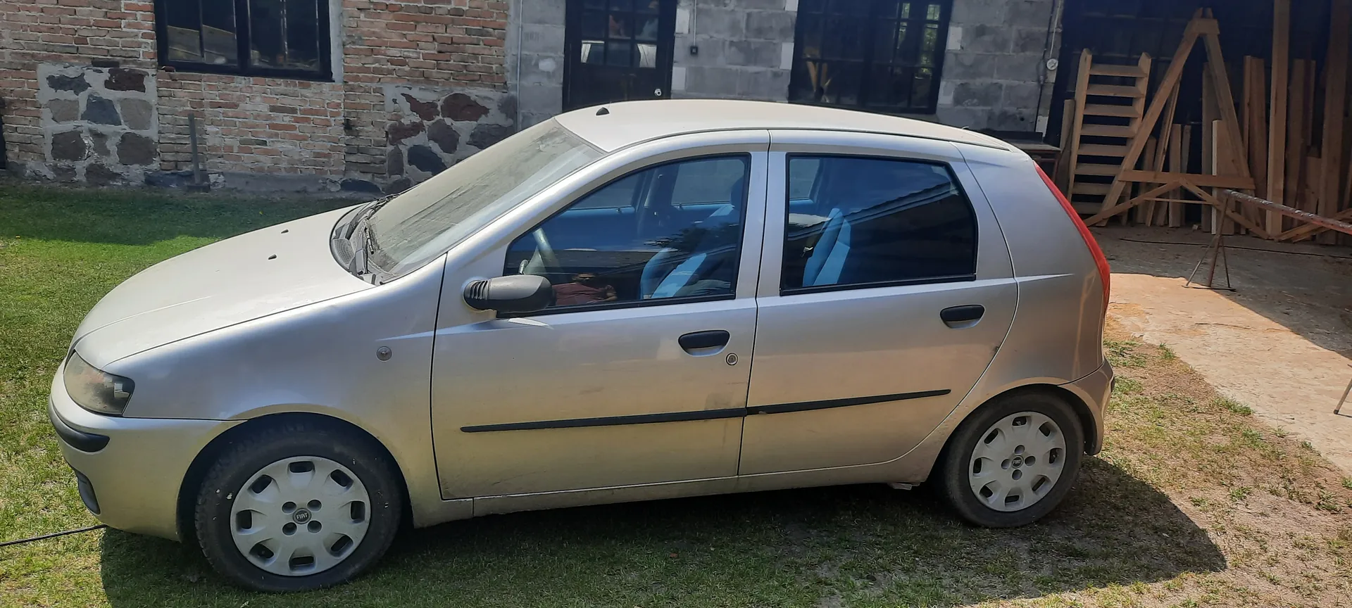 Srebrny samochód hatchback zaparkowany na trawie przed budynkiem z ceglaną i kamieniczną ścianą. W tle widoczne są czarne okna, drzwi oraz drewniane deski i drabina po prawej stronie. Na felgach widoczny jest logo Fiat. Samochód ma czarne listwy boczne i czerwone światło tylnego. Trawa jest częściowo wysuszona, a na ziemi widoczne są plamy ziemi.