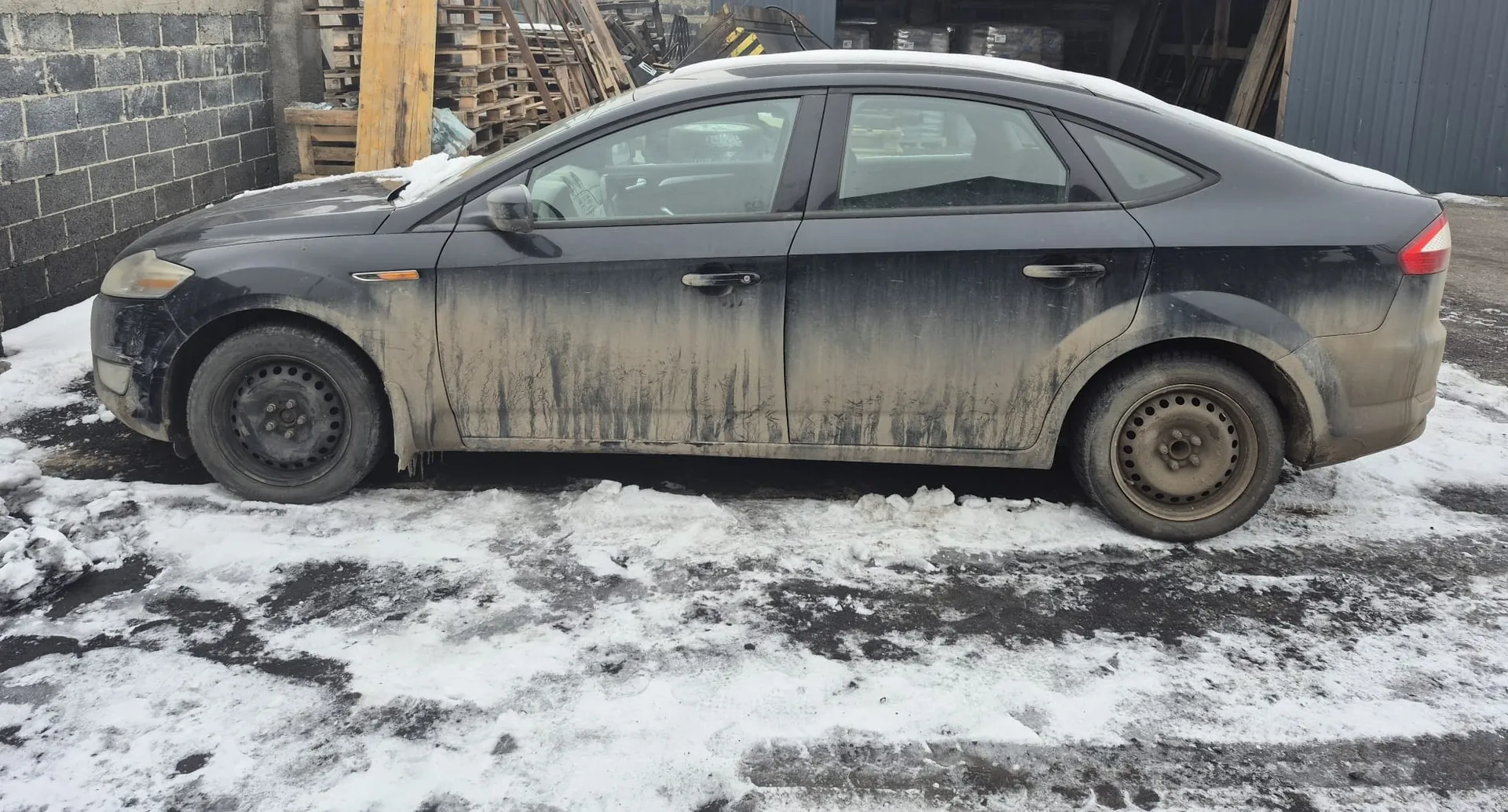 Czarny czterodrzwiowy sedan pokryty grubą warstwą błota, z resztkami śniegu na dachu i masku, zaparkowany na nawierzchni zanieczyszczonej śniegiem i błotem. W tle betonowa ściana, stosy palet i metalowy budynek. Opony zużyte, felgi stalowe bez kapturów.