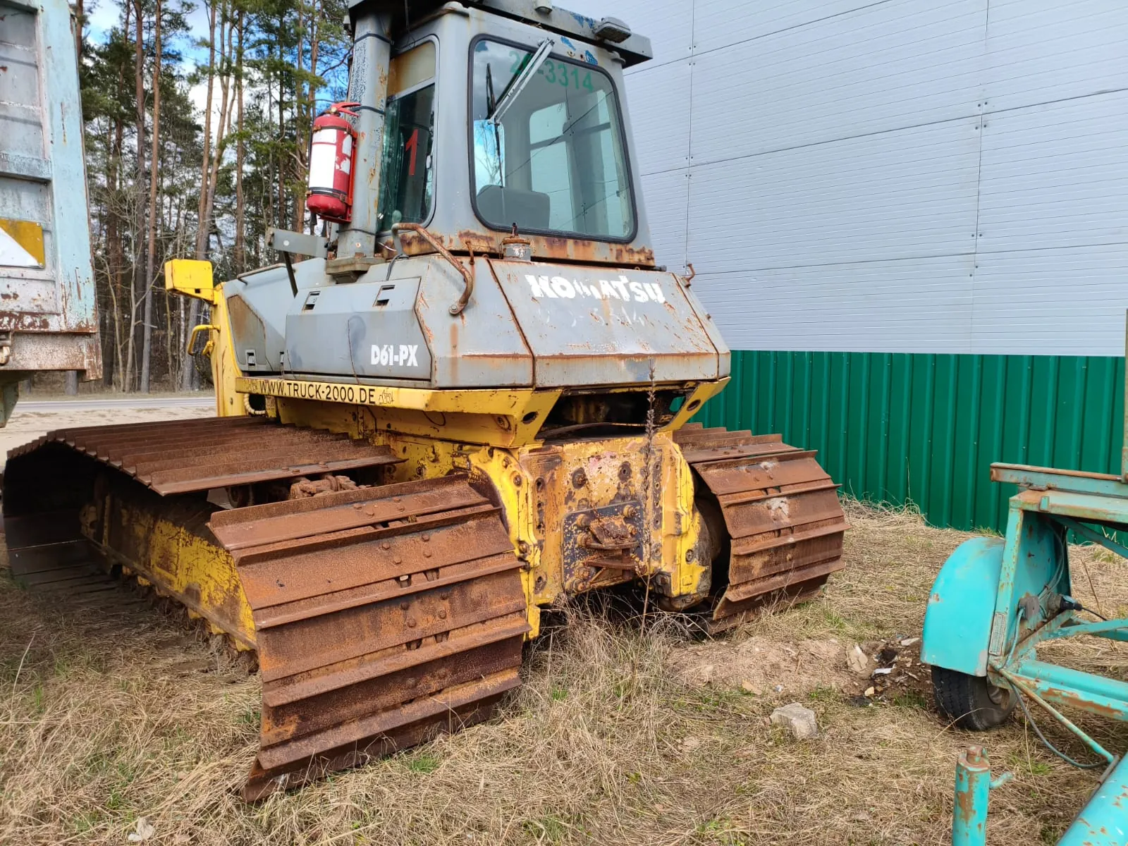 Zardzewiały buldożer gąsienicowy marki Komatsu model D61-PX w kolorze żółto-szarym. Na maszynie widoczne napisy: 'Komatsu', 'D61-PX', 'www.truck-2000.de', numer '1' na szybie kabiny oraz '21-3314' na górze kabiny. Przy maszynie zamontowany czerwony gaśnic. Tło: budynek z poziomymi białymi i zielonymi panelami, sosny, sucha trawa oraz fragmenty innych zardzewiałych maszyn (po lewej i prawej stronie).