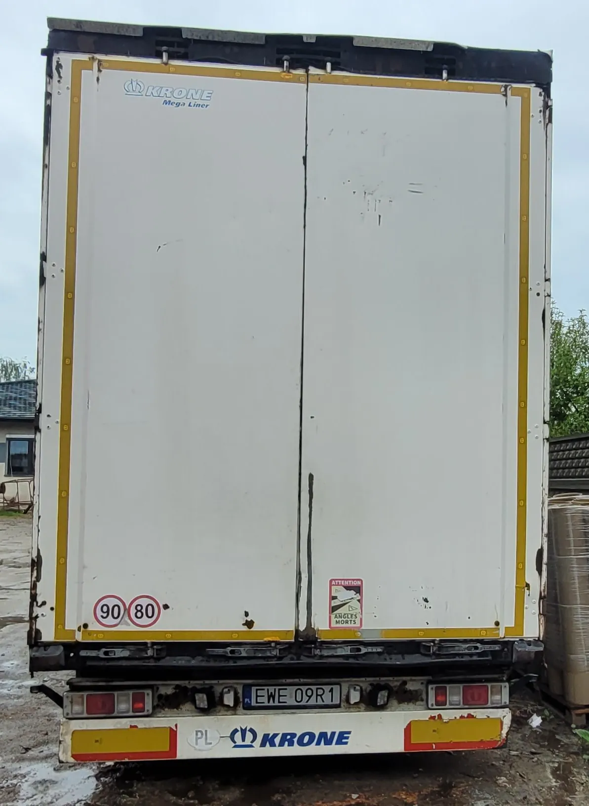 Tylny widok białej naczepy ciężarówki marki Krone Mega Liner. Na białych drzwiach znajdują się okrągłe znaki ograniczenia prędkości 90 i 80 oraz naklejka ostrzegawcza 'ATTENTION ANGLES MORTS'. Na dole znajduje się tablica rejestracyjna o numerze EWE 09R1 oraz napis 'PL KRONE' na czerwono-żółtym pasie odblaskowym.