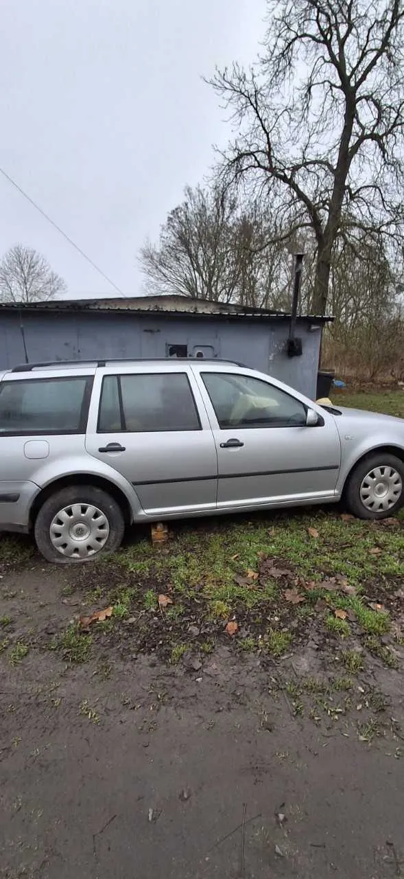 Srebrny samochód typu kombi zaparkowany na błotnistym terenie z trawą i suchymi liśćmi. W tle szara budowla z dachem, kominem oraz drzewami bez liści. Niebo jest pochmurne. Samochód stoi na podkładce pod przednim kołem po lewej stronie.
