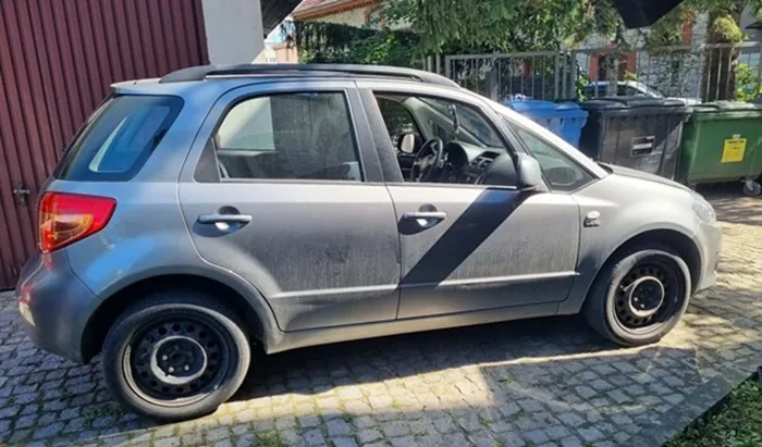 Szary samochód osobowy typu hatchback zaparkowany bokiem na kostce brukowej. Pojazd jest zakurzony i posiada czarne stalowe felgi. W tle widoczna jest czerwona brama, zieleń oraz kilka dużych kontenerów na odpady w kolorach niebieskim, czarnym i zielonym.
