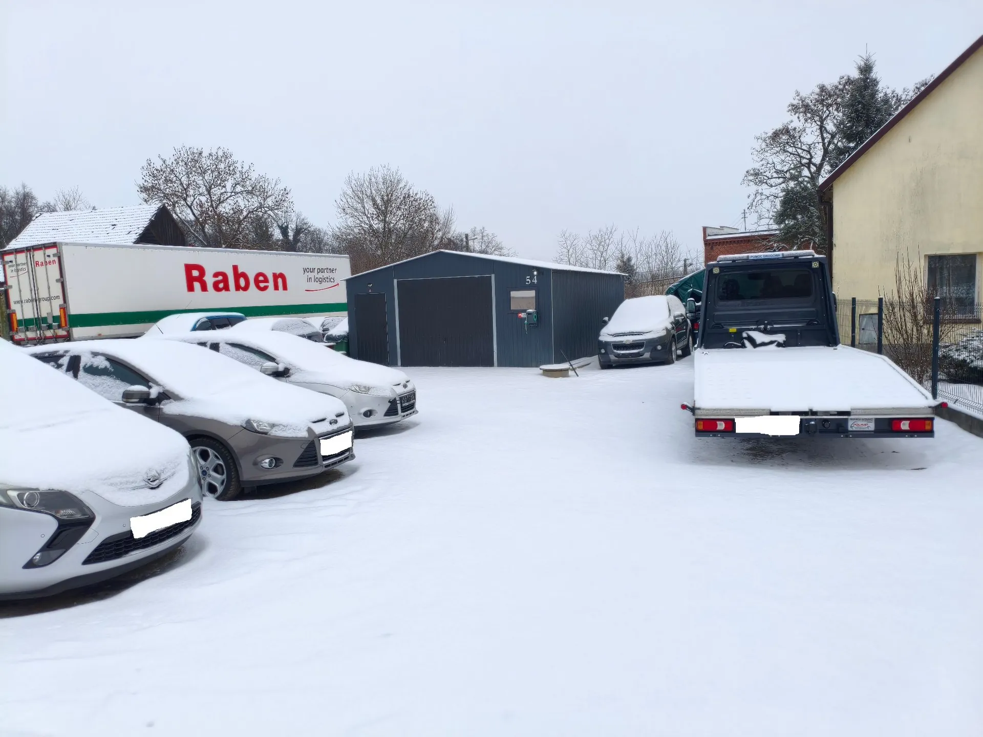 Zasypany śniegiem parking z kilkoma samochodami osobowymi, ciężarówką oraz lawetą. Po lewej stronie stoi biała naczepa z czerwonym napisem 'Raben' i mniejszym tekstem 'your partner in logistics'. W tle znajduje się szary, metalowy budynek gospodarczy oraz fragment żółtego budynku mieszkalnego po prawej stronie. Niebo jest zachmurzone i białe.