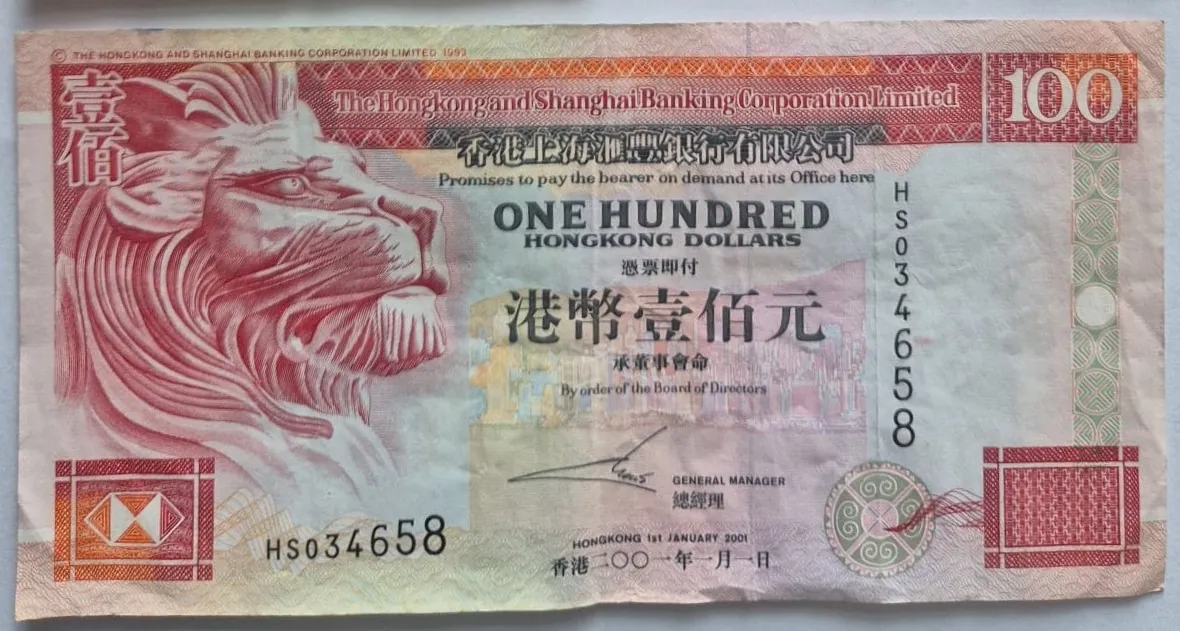 Banknot o nominale 100 dolarów hongkońskich wydany przez The Hongkong and Shanghai Banking Corporation Limited. Po lewej stronie znajduje się duży, czerwony profil lwa. W centralnej części widnieje napis 'ONE HUNDRED HONGKONG DOLLARS' oraz tekst w języku chińskim. W prawym górnym rogu widoczna jest liczba '100'. Numer seryjny 'HS034658' pojawia się dwukrotnie: pionowo po prawej stronie oraz poziomo w dolnej części banknotu. Na dole widnieje data '1st JANUARY 2001' oraz podpis General Managera.