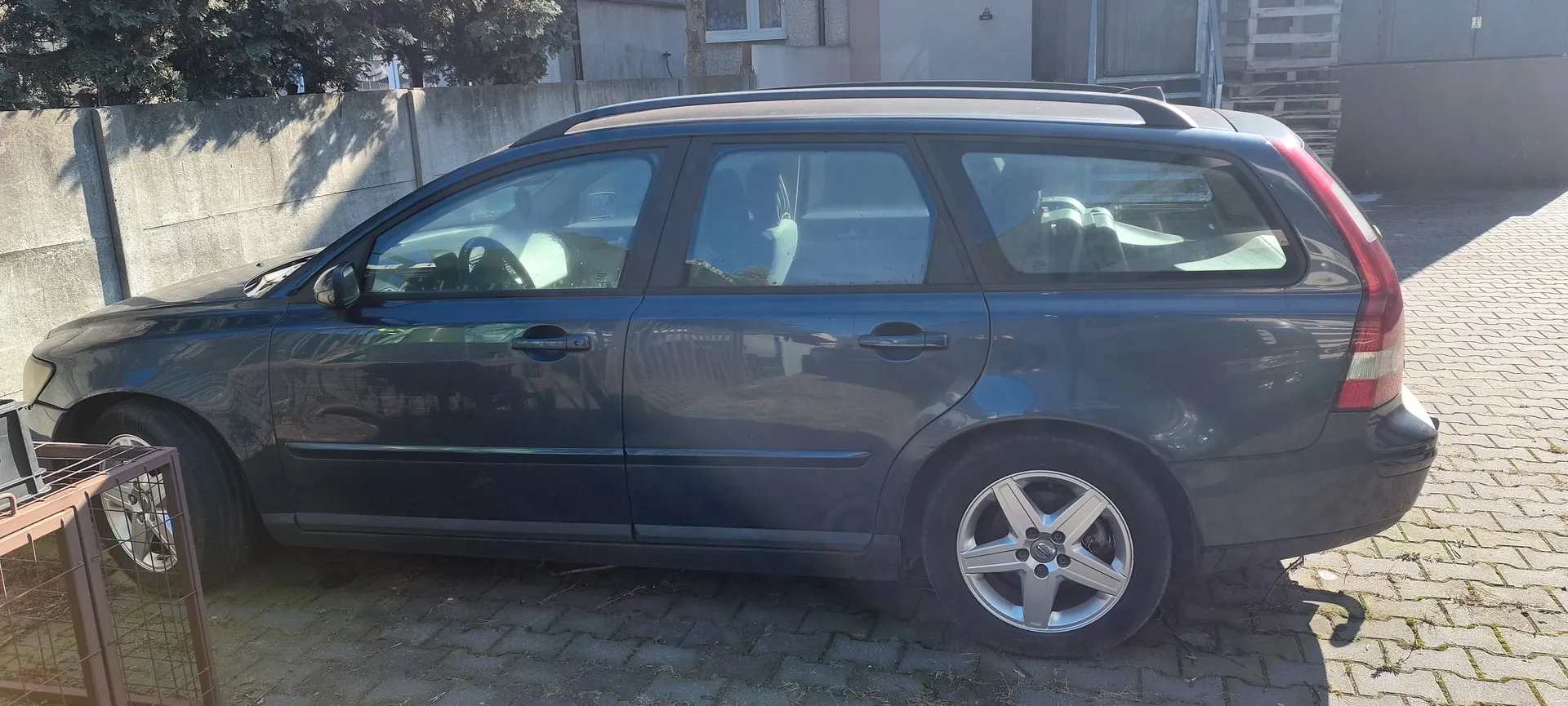 Ciemnoszary samochód typu kombi zaparkowany na brukowanej powierzchni. Po lewej stronie widoczna betonowa ściana, drzewa i metalowa konstrukcja. W tle budynek z oknami i drzwiami. Samochód posiada dachówkę i srebrne felgi. Słońce rzuca cienie na powierzchnię.