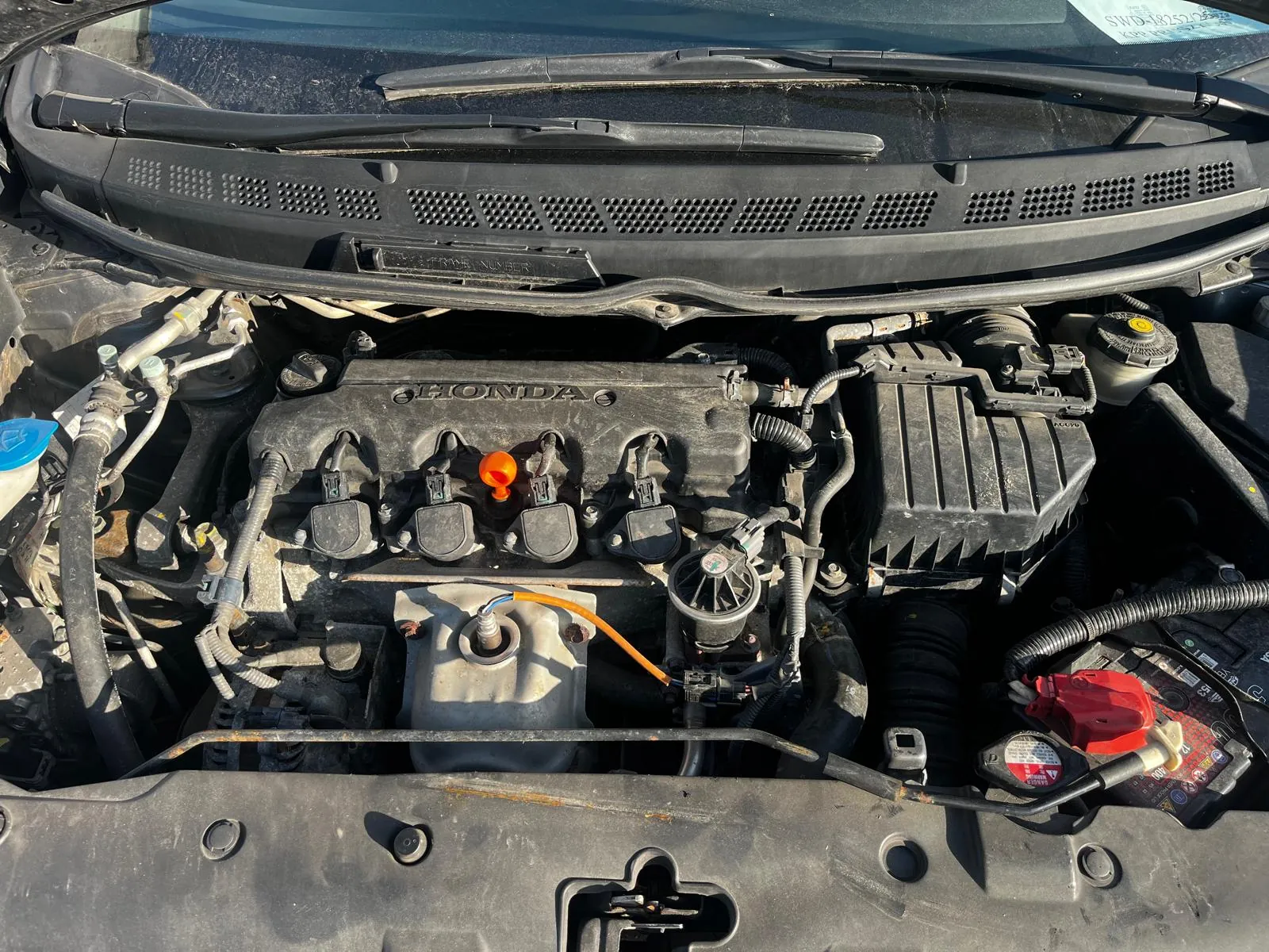 Silnik samochodu marki Honda z otwartą maską. Widoczne są elementy takie jak pomarańczowa nakrętka, niebieska nakrętka, czerwona nakrętka, przewody, węże oraz zbiornik powietrza. Na przedniej szybie widoczny napis SWD-18252. Wycieraczki i siatka wentylacyjna na klapie silnika są również widoczne.