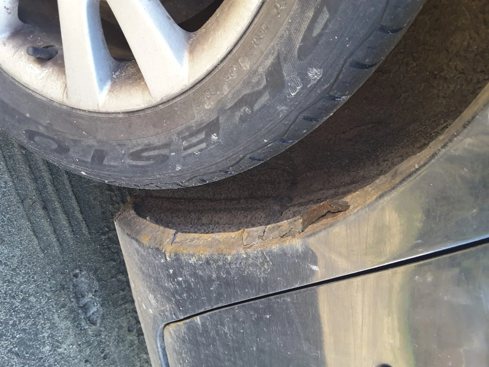 Zbliżenie na koło samochodu i przylegającą część nadwozia. Opona czarna z widocznym napisem 'EST' oraz innymi oznaczeniami. Nadwozie w okolicy otworu kołowego pokryte jest rdzą i uszkodzone, z widocznymi pęknięciami i korozją. Podłoga asfaltowa. Nadwozie ciemne, pokryte brudem.