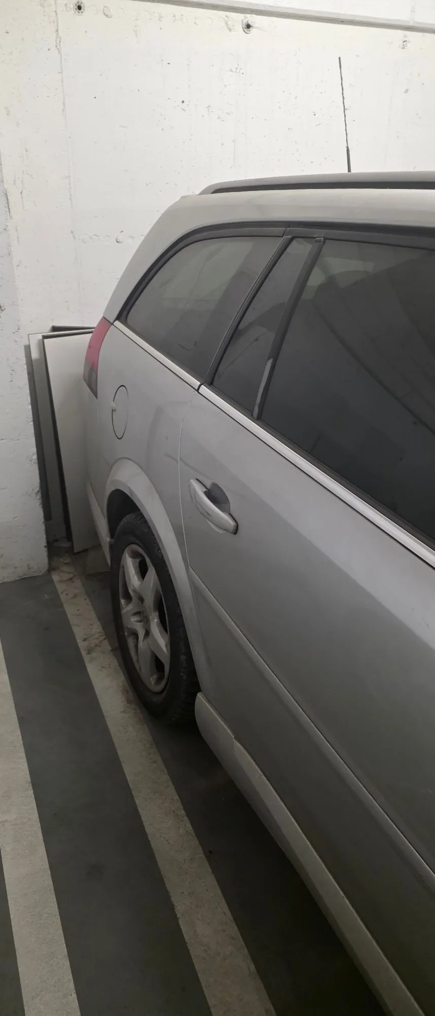 Boczny widok srebrnego samochodu typu kombi zaparkowanego w garażu obok białej ściany. Widoczna jest tylna część nadwozia, koło oraz przyciemniane szyby.