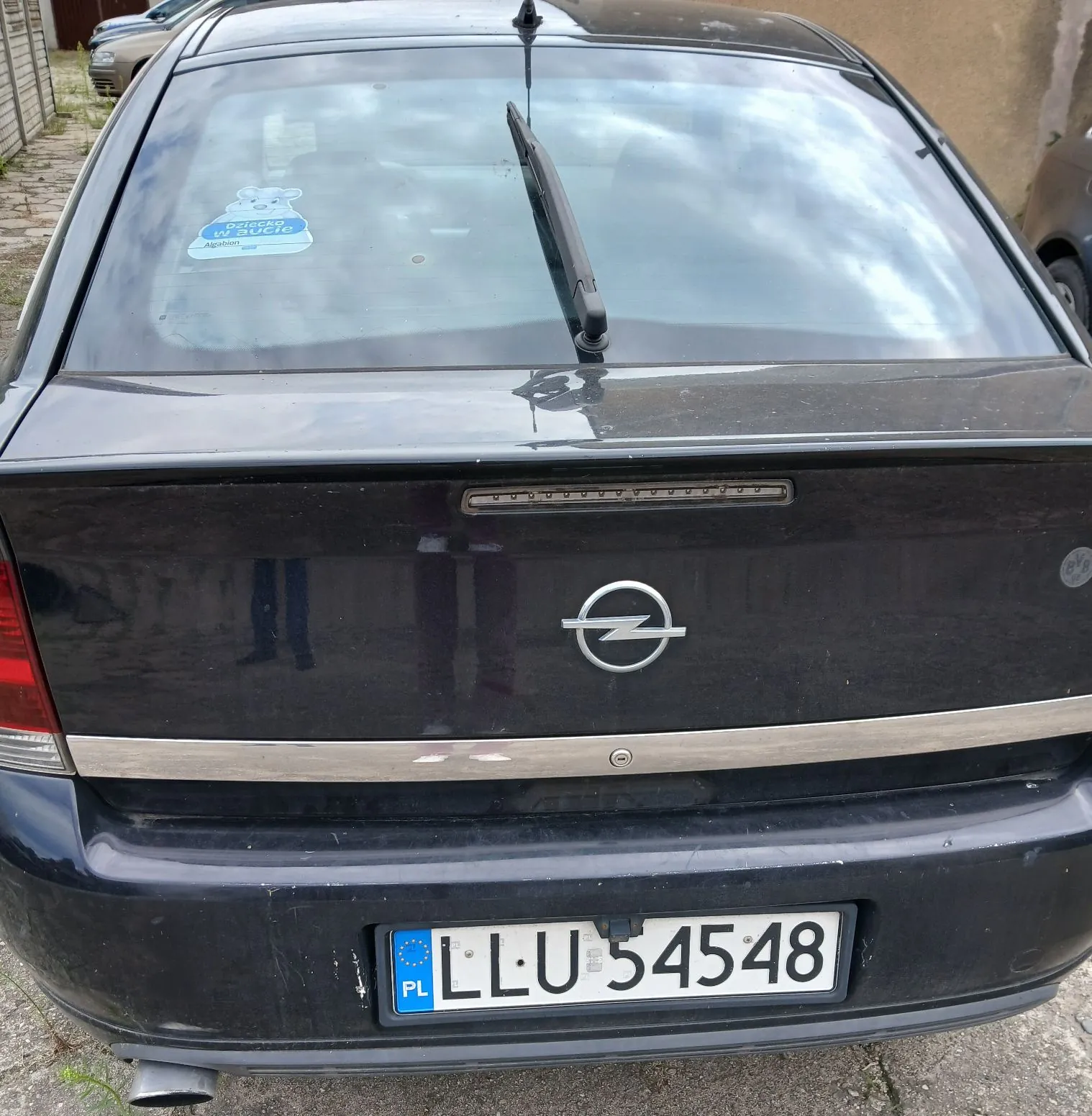 Tylny widok czarnego samochodu marki Opel. Na tylnej szybie znajduje się niebieska naklejka z napisem 'dziecko w aucie'. Na klapie bagażnika widoczne jest logo Opela oraz chromowana listwa. Samochód posiada polską tablicę rejestracyjną o numerze LLU 54548.