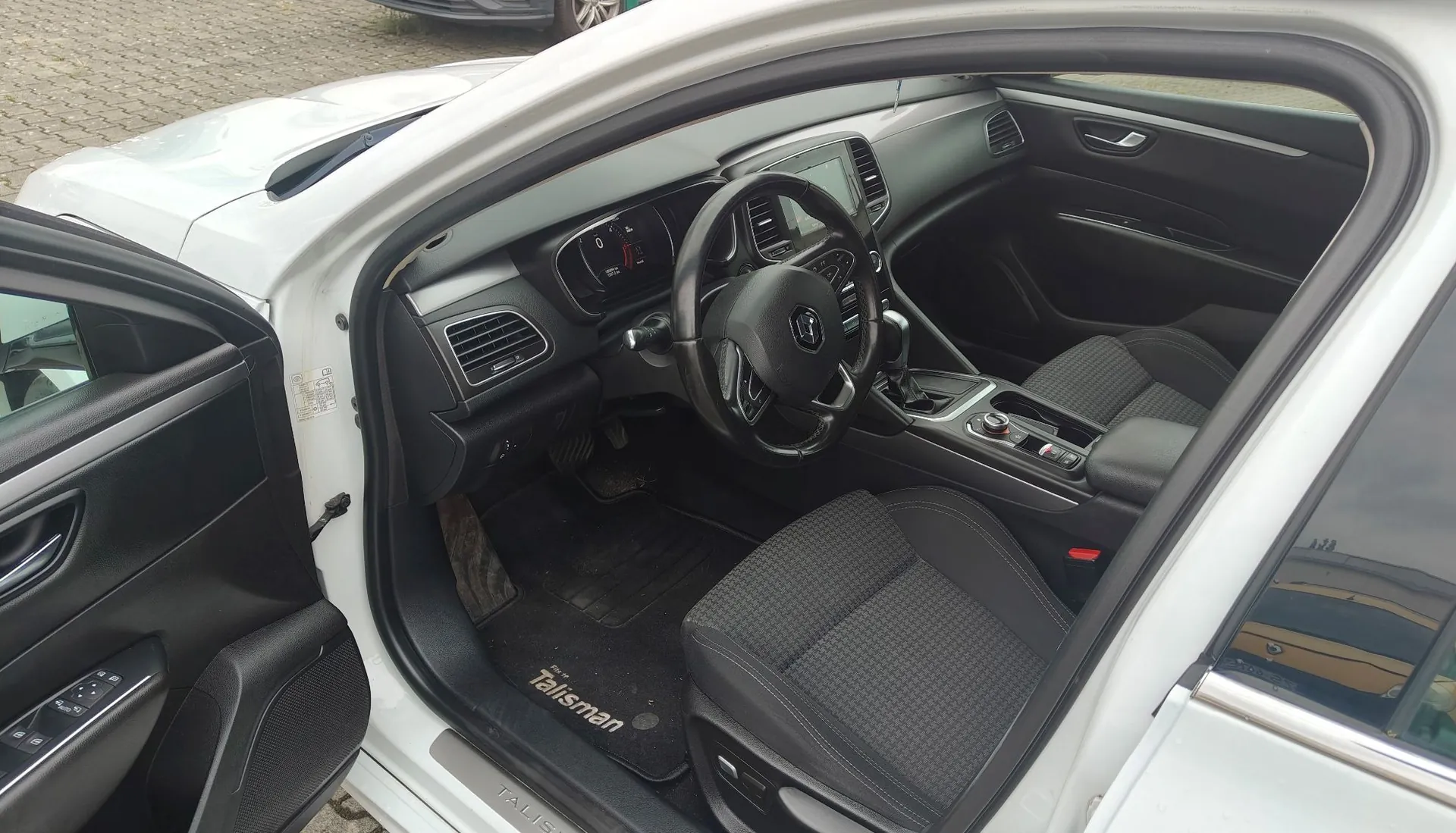 Wnętrze białego samochodu marki Renault Talisman widziane z otwartych drzwi kierowcy. Widoczna jest czarna tapicerka foteli, kierownica, deska rozdzielcza z ekranem i zegarami oraz dywanik z napisem 'Talisman'.