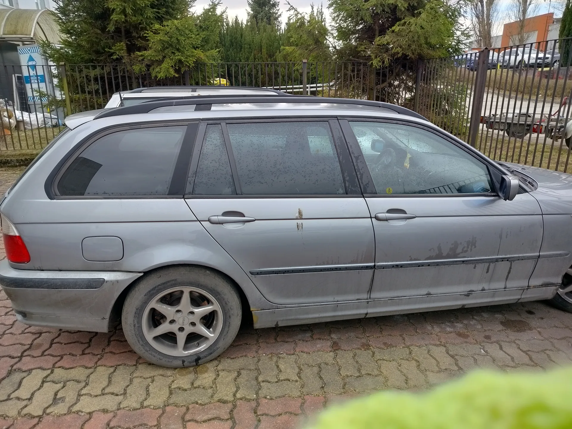 Srebrny samochód typu kombi zaparkowany na brukowanej powierzchni. Pojazd ma zabrudzoną karoserię z widocznymi plamami błota i rdzy w okolicach klamek drzwi. Tylne okno i boczne szyby są przyciemniane, na przedniej szybie pasażera widoczny żółty naklejka. W tle metalowe ogrodzenie, drzewa iglaste, fragment budynku z niebieskim znakiem oraz inne zaparkowane pojazdy. Niebo jest zachmurzone.