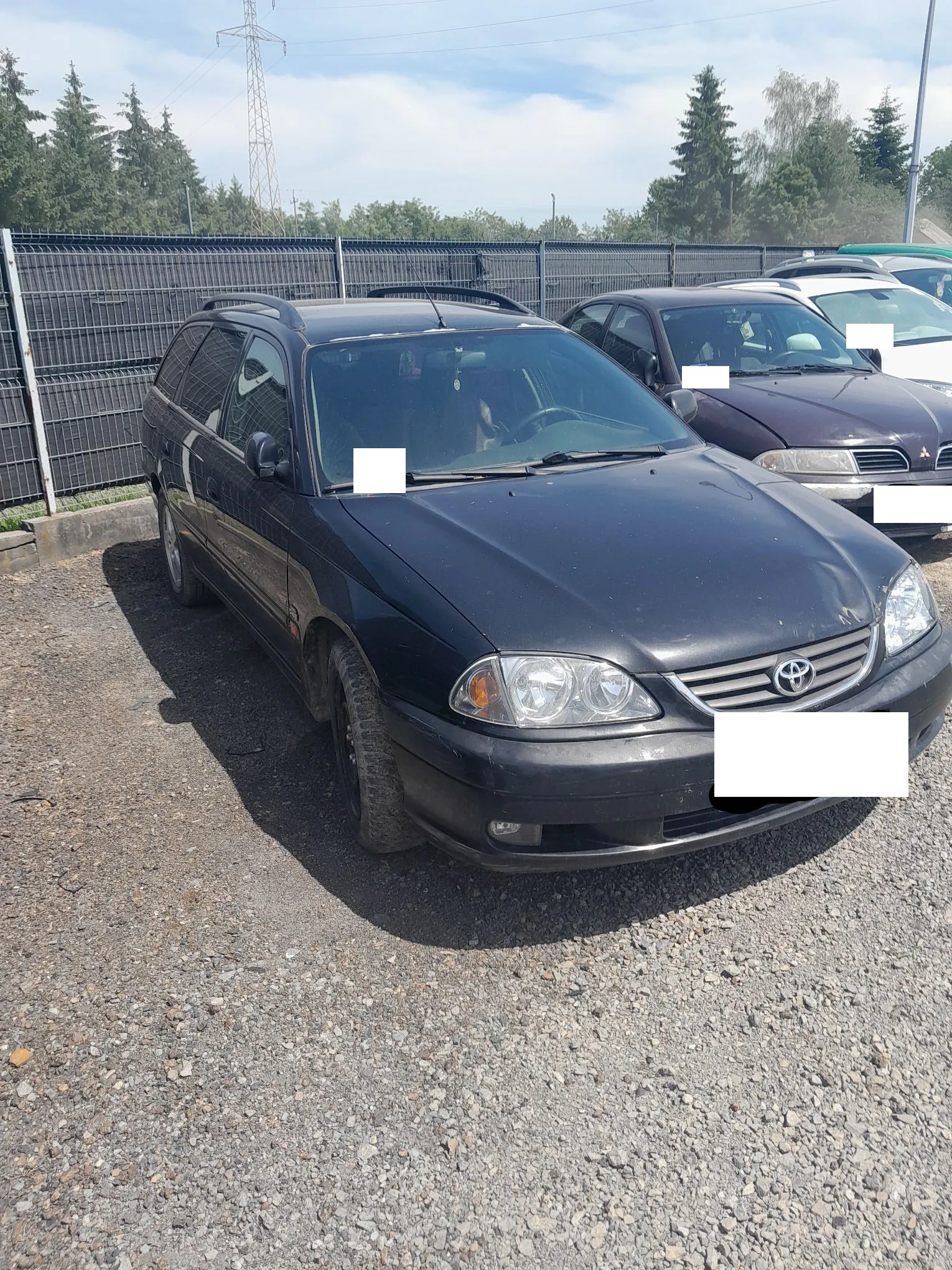 Czarny samochód marki Toyota w nadwoziu kombi zaparkowany na żwirowym placu. Auto jest widoczne z przodu i z boku. W tle znajduje się metalowy płot, drzewa oraz jasne niebo z lekkimi chmurami. Obok stoi inny, ciemnofioletowy samochód. Tablice rejestracyjne oraz niektóre detale są zakryte białymi prostokątami.