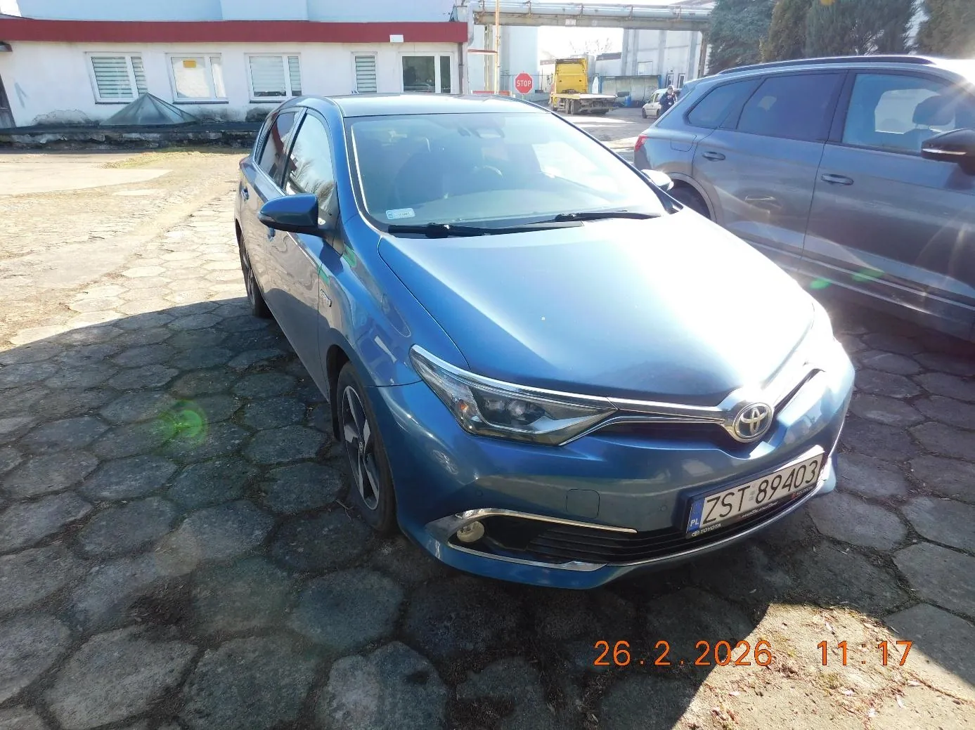 Niebieski samochód marki Toyota z tablicą rejestracyjną ZST 89403 zaparkowany na brukowanej powierzchni. Obok niego stoi szary samochód typu SUV. W tle budynek z białymi ścianami i czerwonym pasem, oknami z żaluzjami oraz czerwony znak STOP. Widoczny również żółty pojazd ciężarowy i drzewa. Na dole zdjęcia pomarańczowy znaczek z datą 26.2.2026 i godziną 11:17.