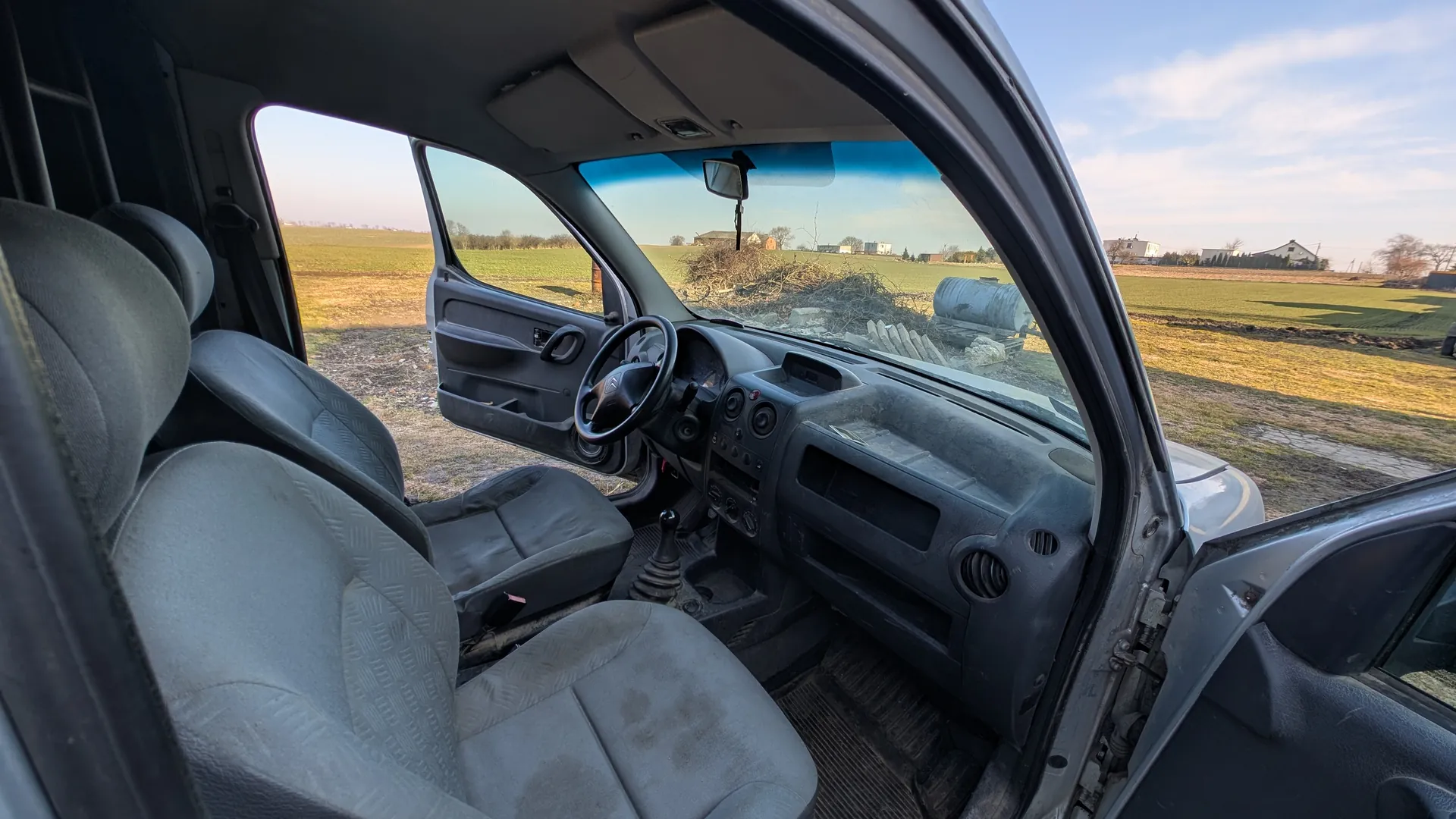 Wnętrze samochodu z otwartymi drzwiami od strony kierowcy i pasażera. Widoczne są szare siedzenia z wzorem, kierownica z logo, zatłuszczona deska rozdzielcza z przyciskami i wskaźnikami, dźwignia zmiany biegów oraz podłoga z brudnymi dywanikami. Poza samochodem rozciąga się pole, widoczne są budynki, duży metalowy zbiornik oraz sterta gałęzi. Niebo jest częściowo zachmurzone.