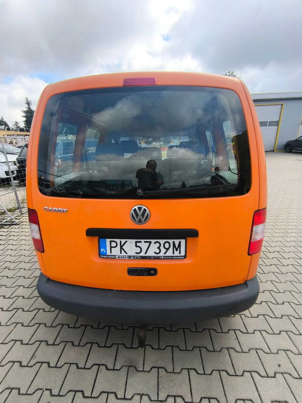 Tylny widok pomarańczowego samochodu dostawczego marki Volkswagen Caddy. Pojazd posiada tablicę rejestracyjną o numerze PK 5739M z oznaczeniem PL. Pod tablicą znajduje się napis: 'SERWIS WARSZAWA 507...'. Samochód stoi na kostce brukowej na tle zachmurzonego nieba i budynku przemysłowego.