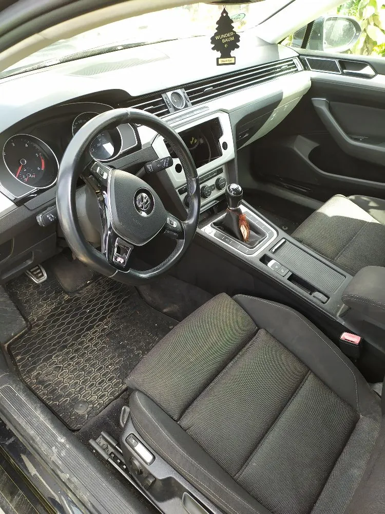 Wnętrze samochodu marki Volkswagen widziane z perspektywy kierowcy. Widoczna jest czarna kierownica z logo marki, deska rozdzielcza z ekranem multimedialnym, zegary analogowe oraz ciemnoszare, materiałowe fotele. Na lusterku wstecznym wisi zapach samochodowy w kształcie choinki z napisem 'WUNDER-BAUM'. Podłoga wyłożona jest gumowymi dywanikami o strukturze plastra miodu.