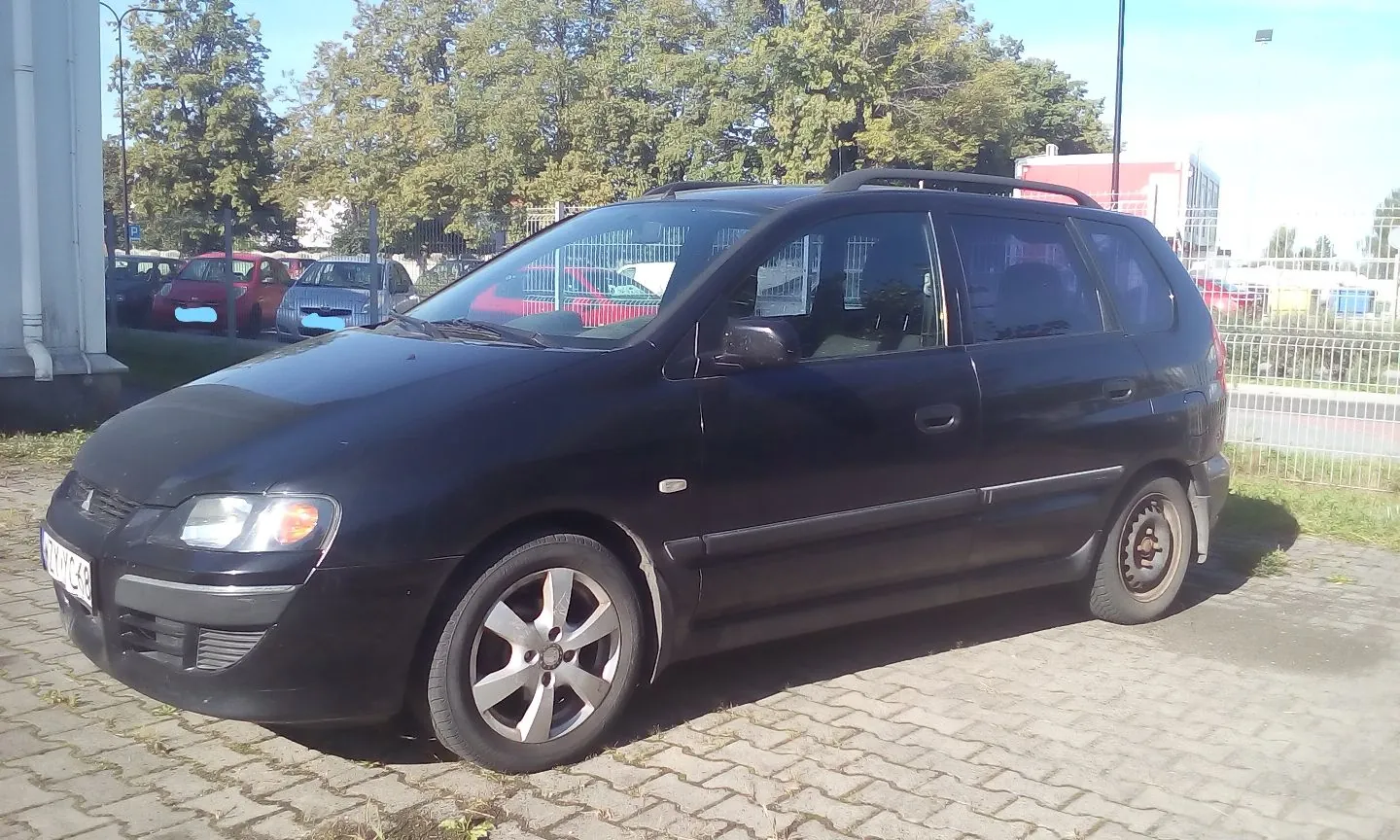 Czarny samochód osobowy typu kombi zaparkowany na kostce brukowej. Pojazd jest ustawiony bokiem do obiektywu, z przodu widoczny jest reflektor i fragment tablicy rejestracyjnej. W tle znajdują się inne zaparkowane samochody, zielone drzewa oraz biały ogrodzenie z siatki.