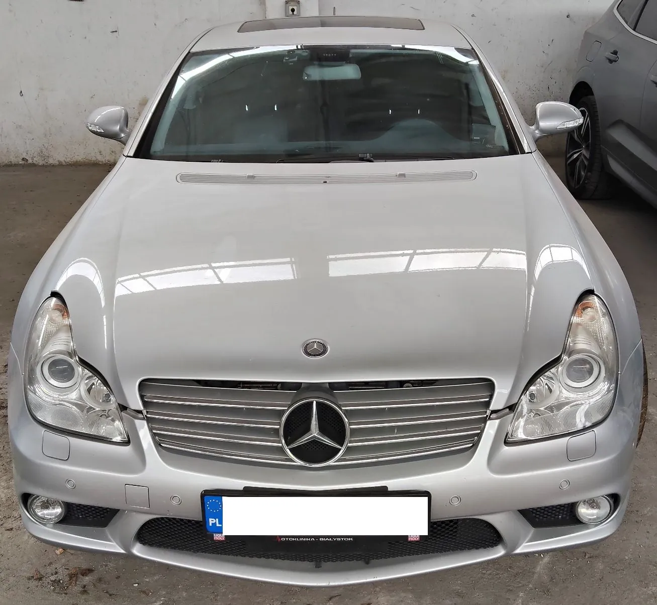 Srebrny samochód marki Mercedes-Benz widziany od przodu. Widoczna jest maska, reflektory, grill z dużym logo gwiazdy Mercedesa oraz zderzak z wbudowanymi światłami przeciwmgielnymi. Samochód posiada białą tablicę rejestracyjną z oznaczeniem PL i logo Unii Europejskiej. Poniżej tablicy znajduje się napis 'OTOKUNKUNKA - BIALYSTOK'.